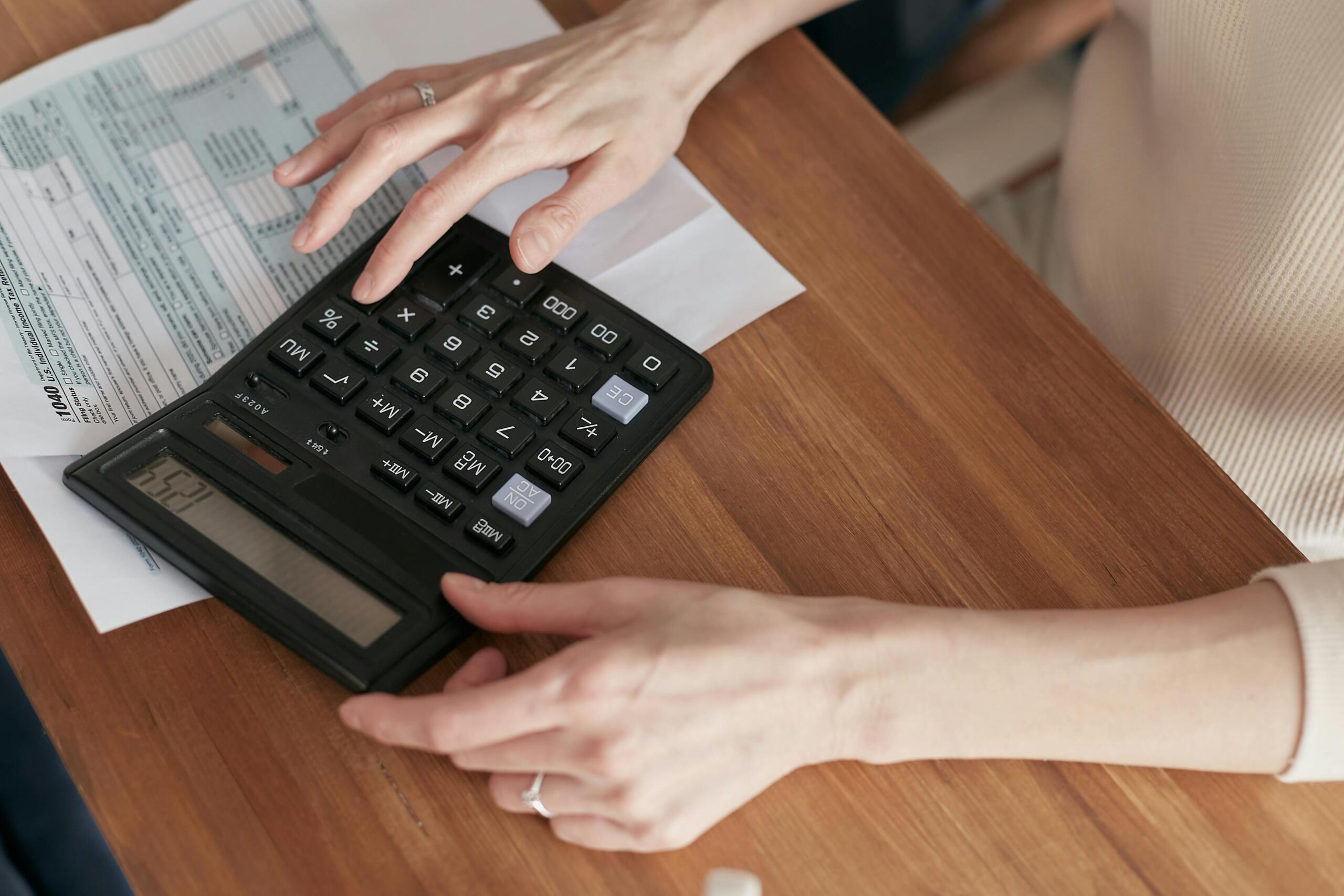 Hands pressing buttons on a large calculator above documents on a wooden table.