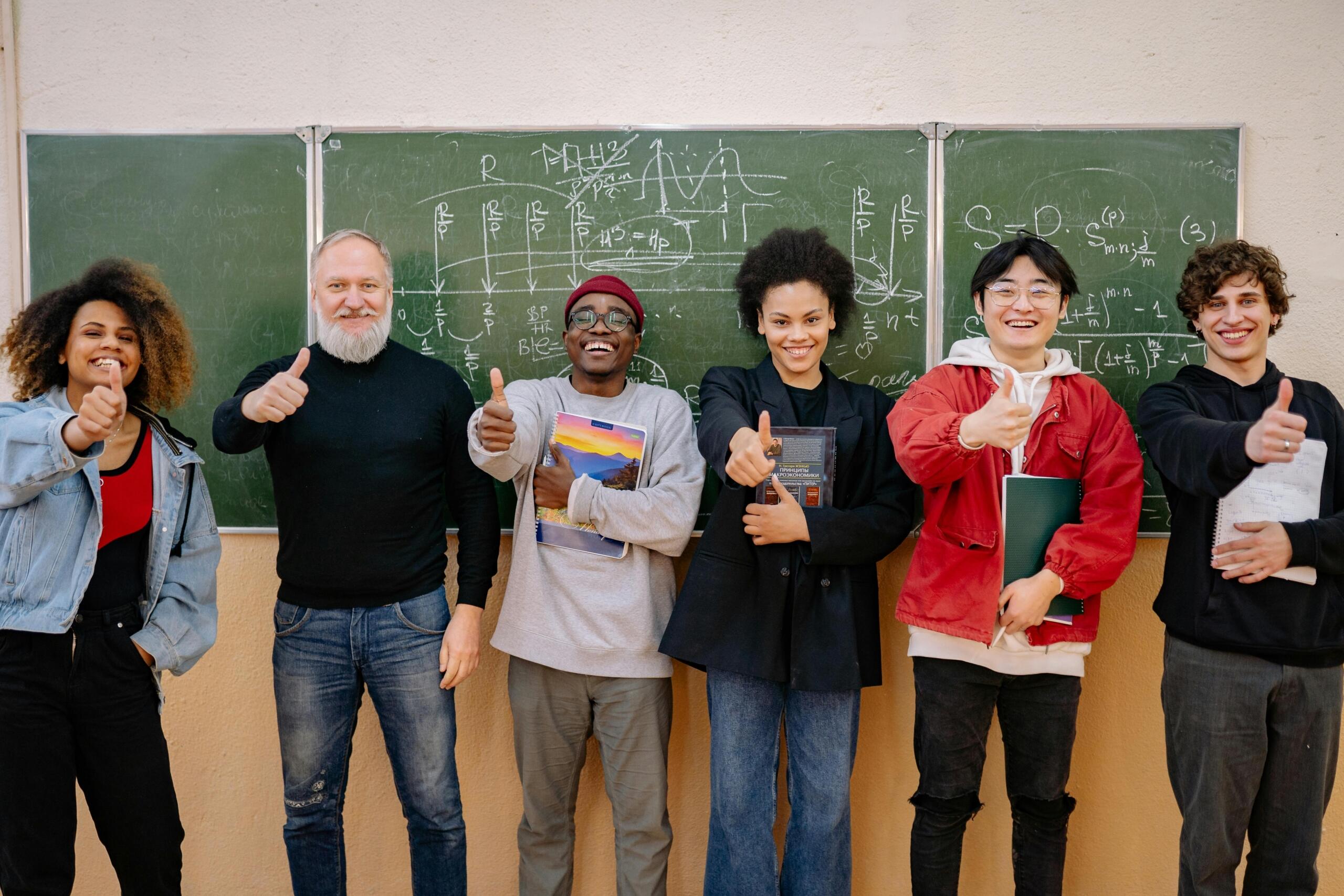 A teacher and students smiling and giving thumbs up in front of a chalkboard with mathematical formulas.