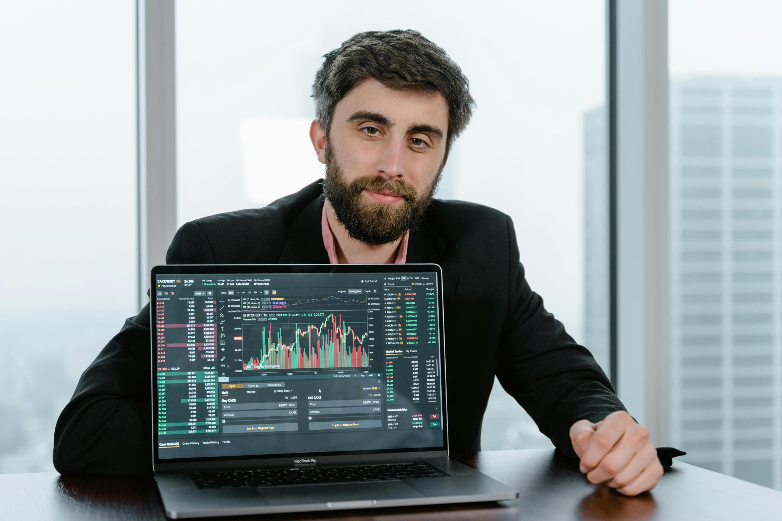 A businessman in a dark coat sits behind a laptop and analyses stock market data on the screen.