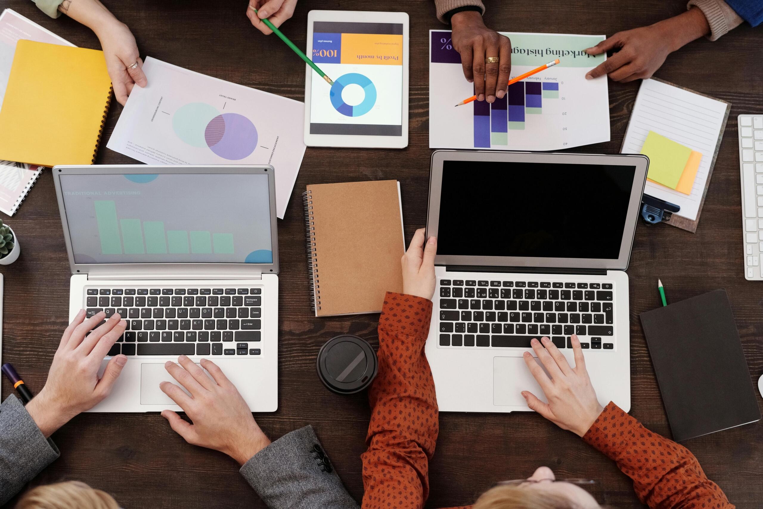 High angle view of multiple people working around a table with two laptops, printed charts and a tablet displaying graphs.