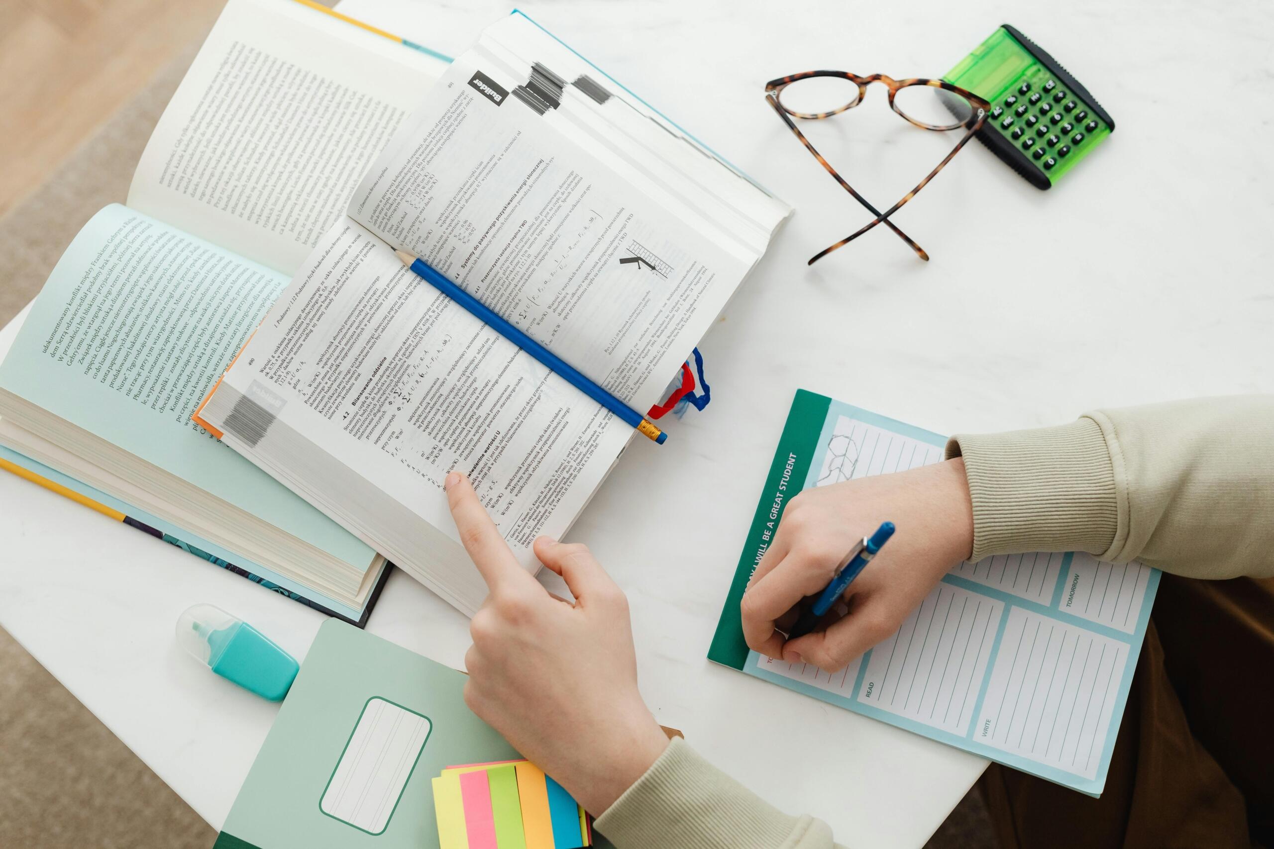 Overhead view of a study desk with open textbooks, a person’s hand pointing at text, a pen in the other hand, glasses, a calculator and a planner.