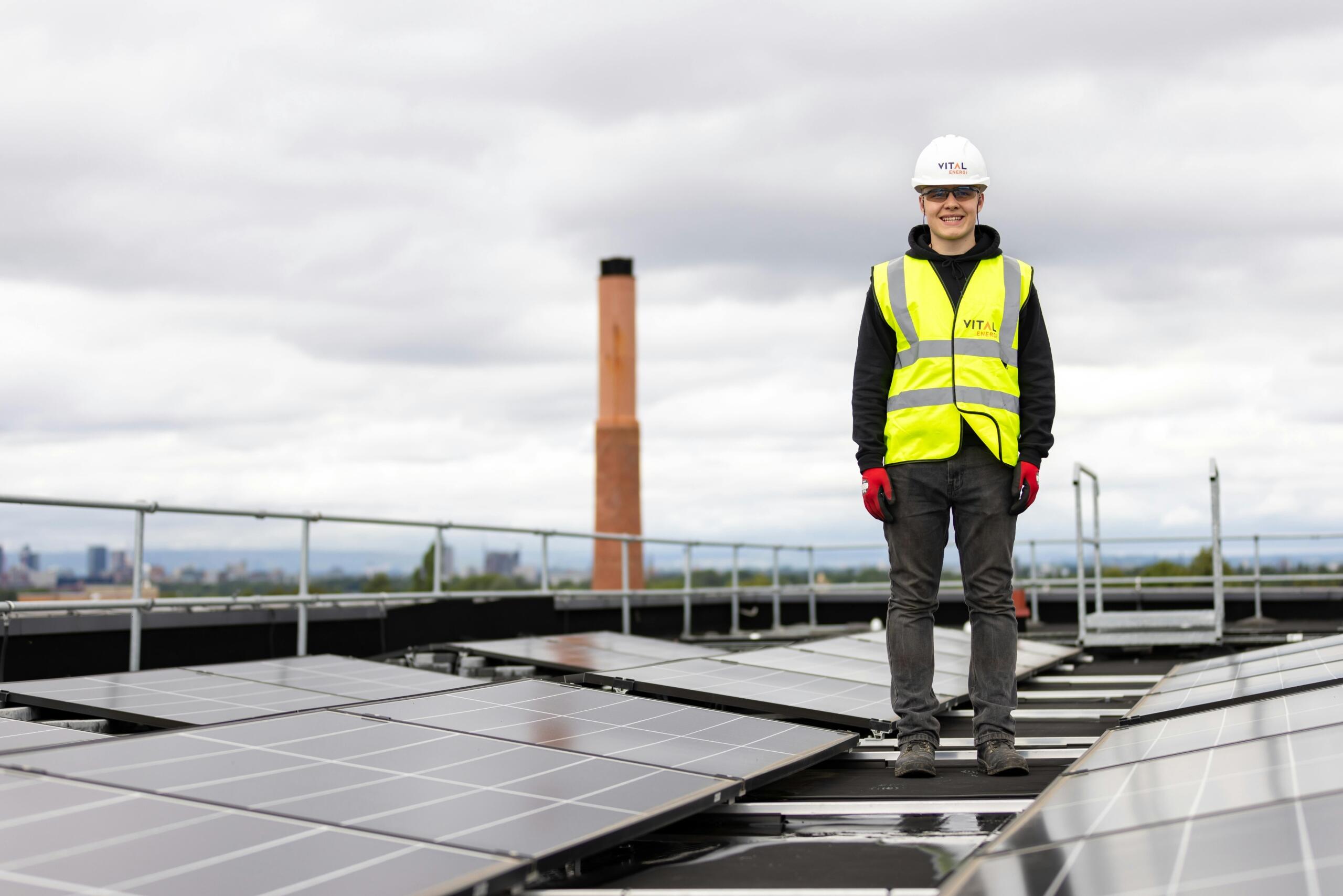 Engineer wearing a high visibility vest and safety helmet standing on a rooftop among solar panels, with a cloudy sky and city skyline in the background.