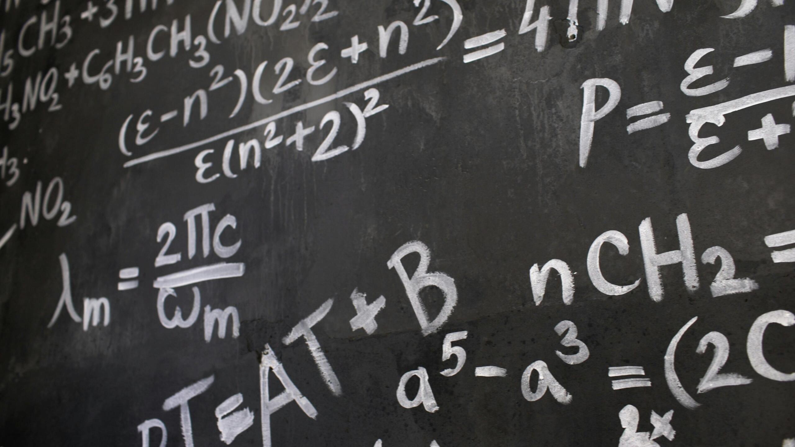 Detailed view of a black chalkboard covered with multiple handwritten algebraic and chemical equations written in white chalk.