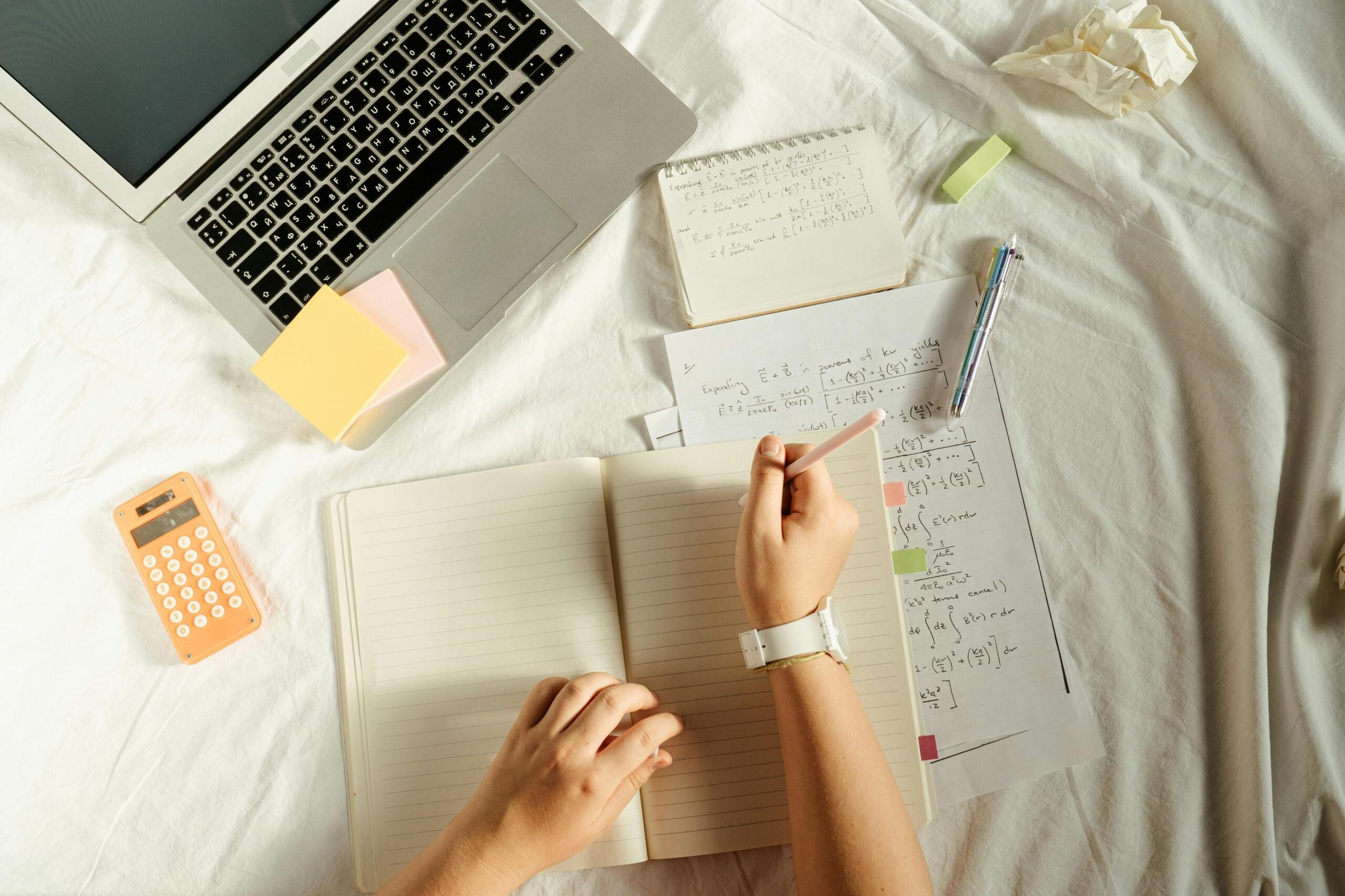 High‑angle photo of a person taking notes in a blank notebook while studying math worksheets beside a laptop, sticky notes and a calculator on a bed.