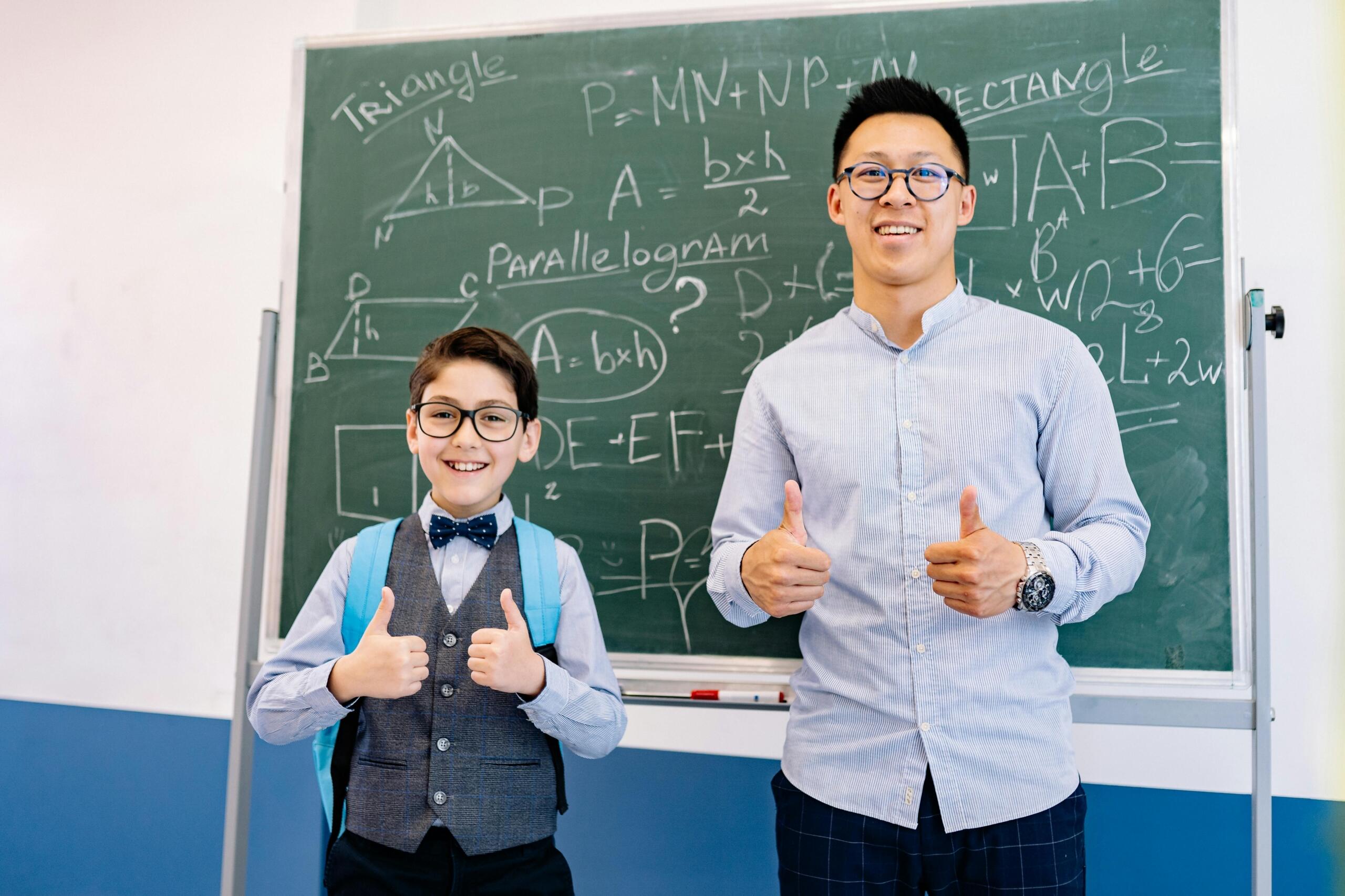 Teacher and young student with glasses standing in front of a chalkboard filled with geometric formulas, both giving thumbs up.