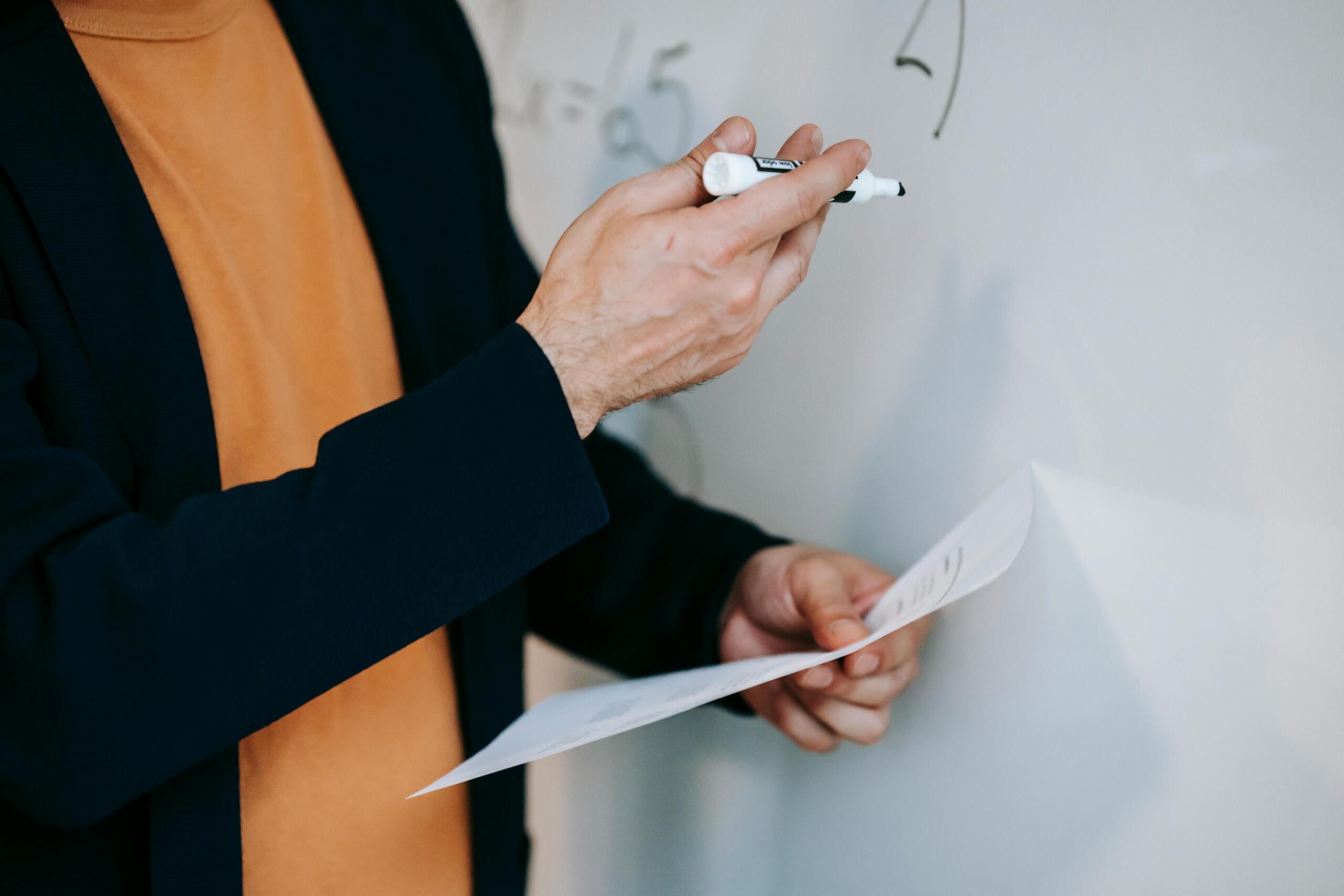 Close up of a teacher holding a whiteboard marker and a sheet of paper while explaining maths on a whiteboard.