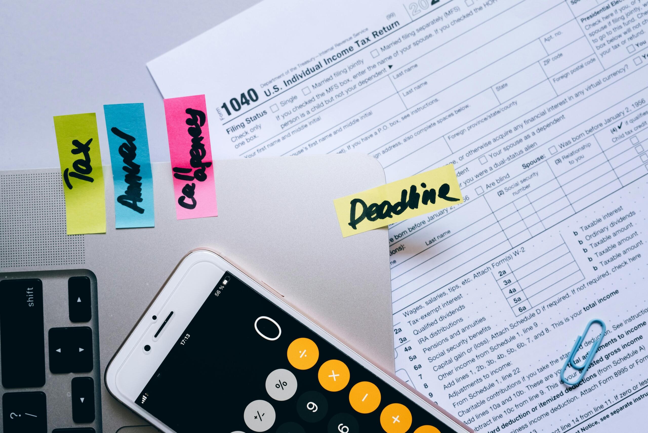 Tax forms, sticky notes, a smartphone calculator and a laptop arranged on a desk.