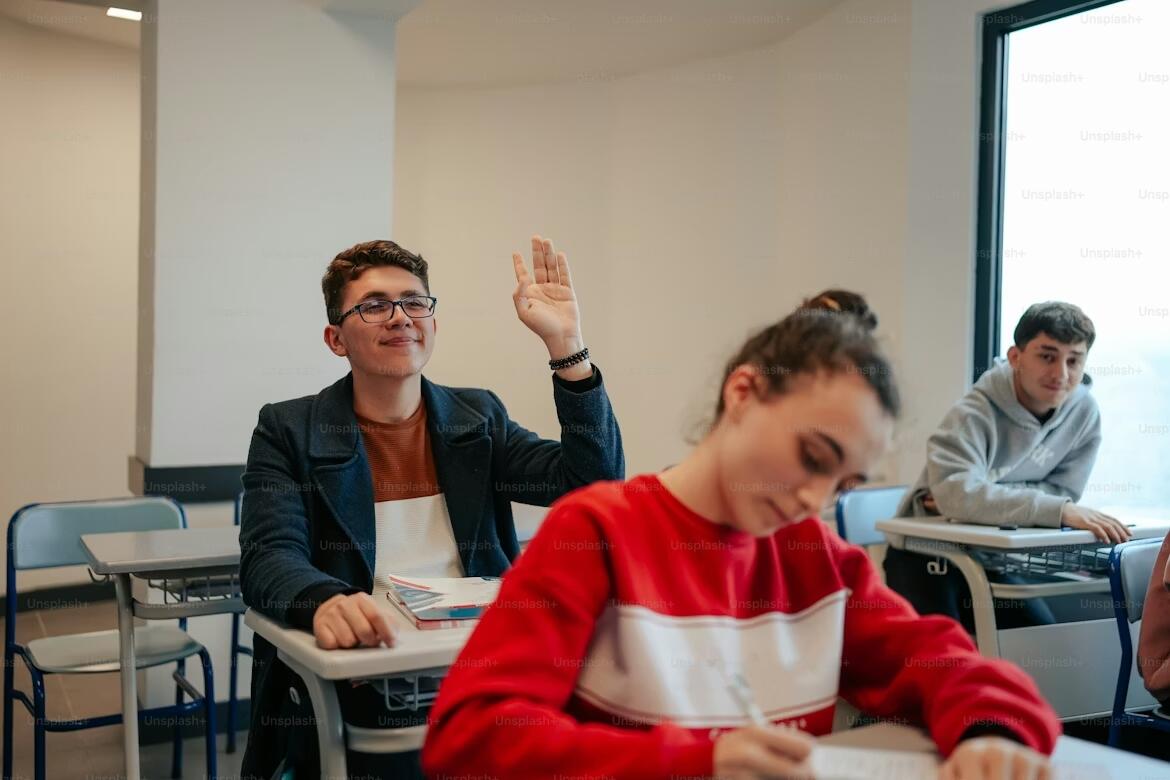 A student raising his hand in a classroom while other students sit at their desks, creating an engaged and interactive learning environment during a language lesson.