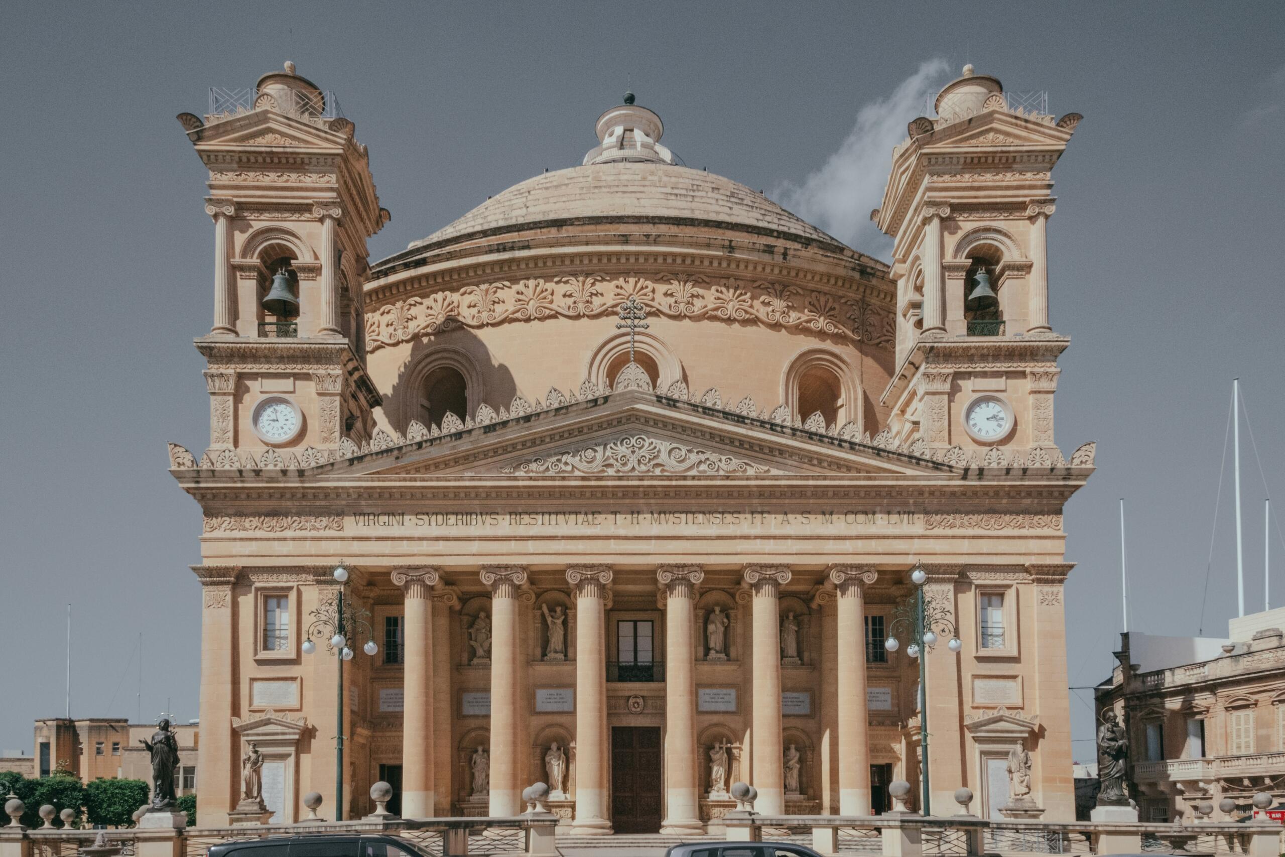 A large limestone church with a central dome, two symmetrical bell towers with clocks, and a columned façade stands against a clear sky.