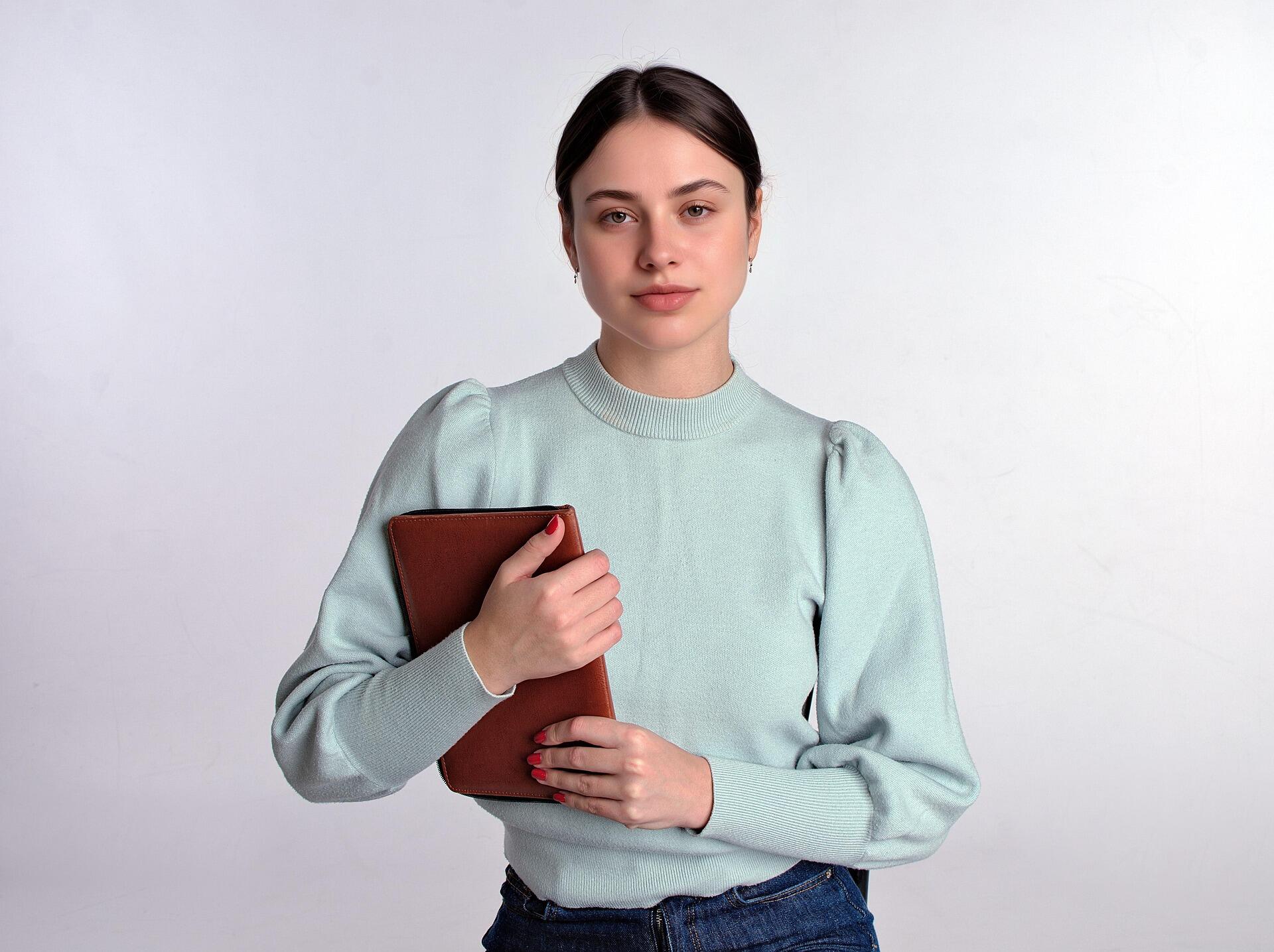 Young woman wearing a light green sweater holding a brown notebook against her chest, standing in front of a plain white background and looking at the camera.