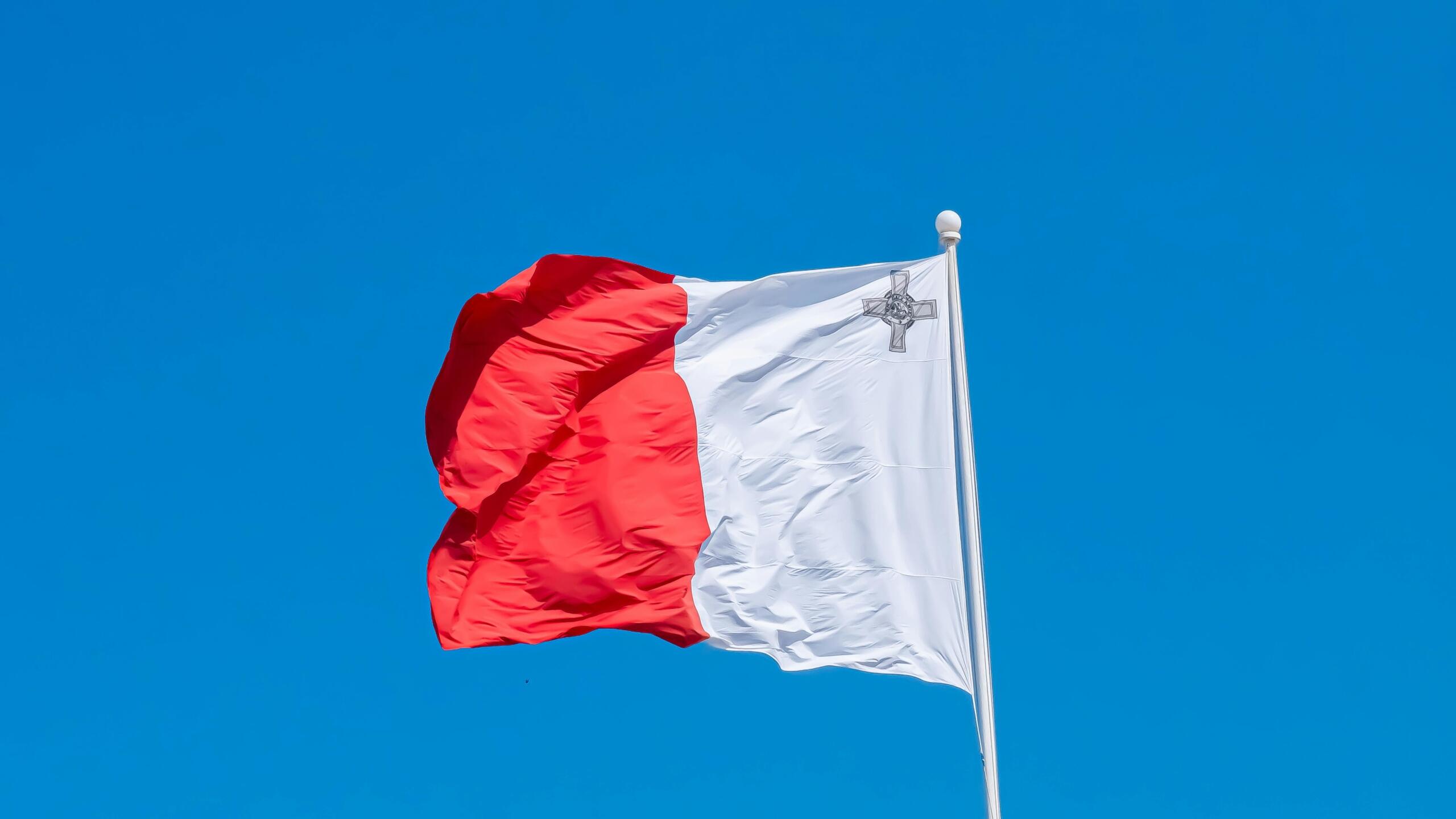 Red and white Maltese flag with the George Cross in the upper corner, waving against a clear blue sky.