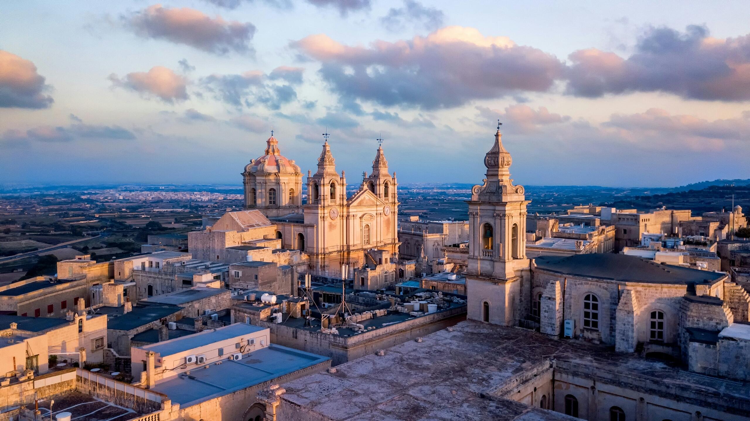 An elevated view shows a limestone cathedral with twin bell towers rising above dense historic buildings, surrounded by countryside under a partly cloudy sky.