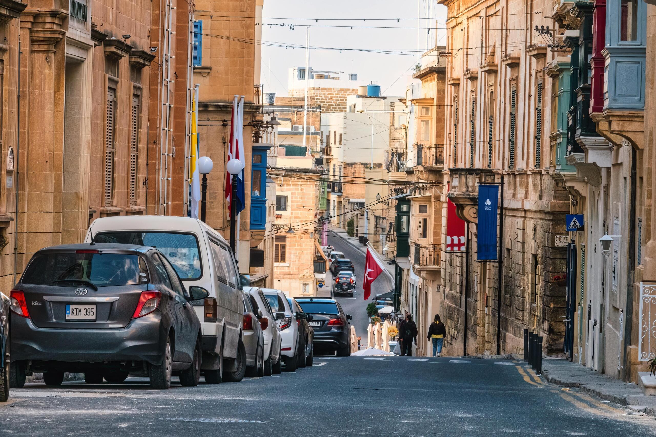 Street in Birgu with limestone facades, parked cars, and Maltese flags hanging across the road.