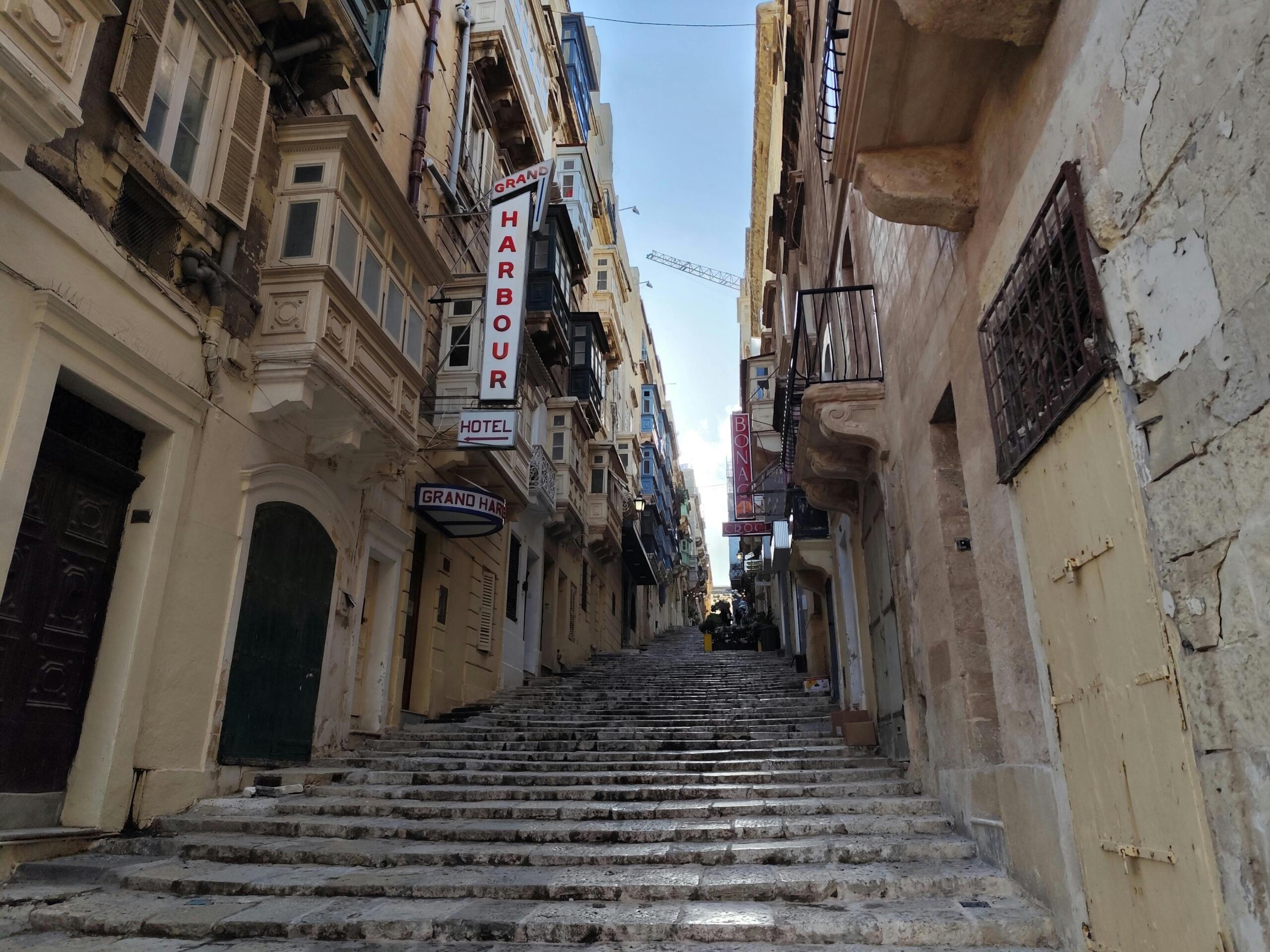 Narrow stepped street in Valletta lined with traditional stone buildings and enclosed wooden balconies in daylight.