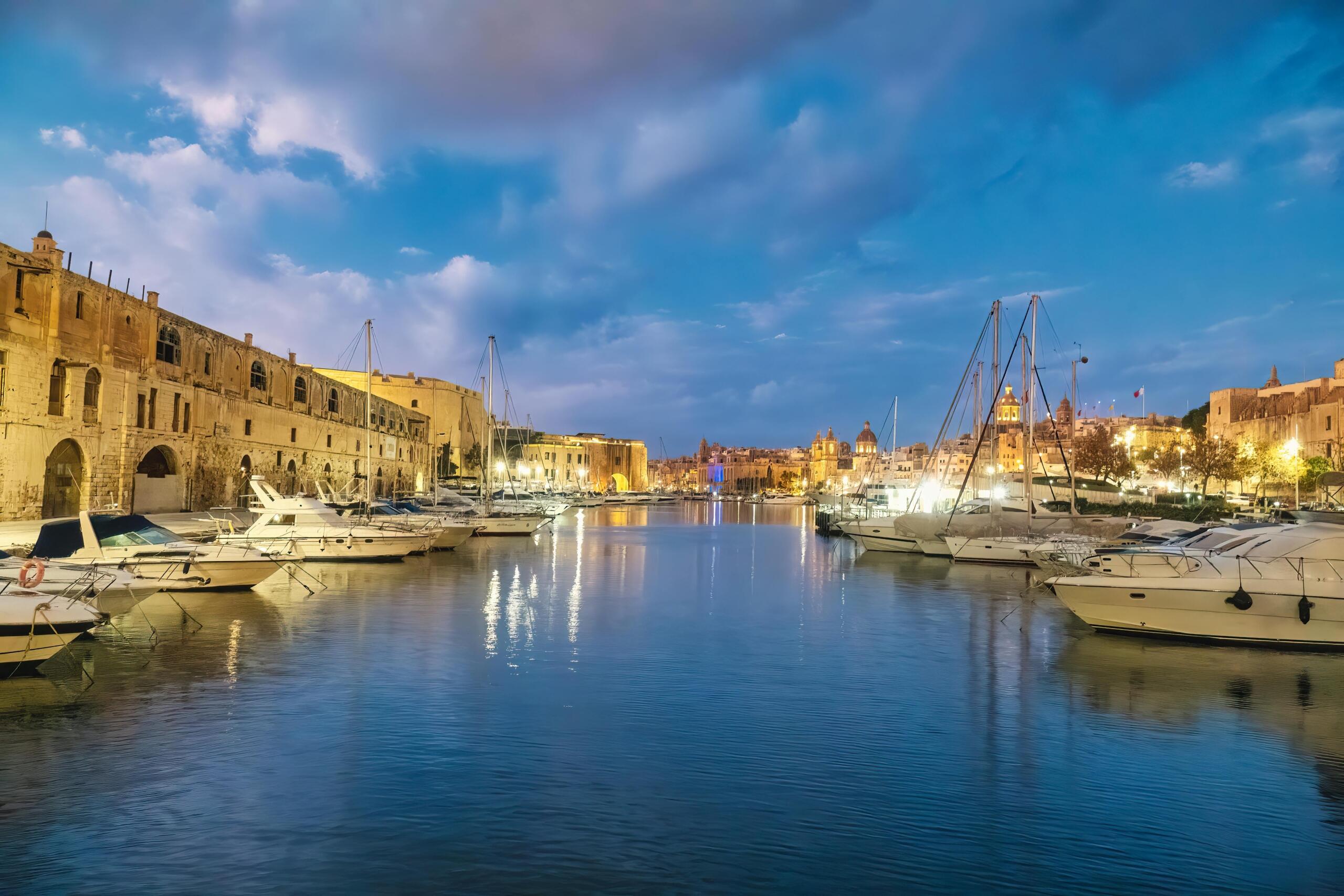 Twilight view over Malta’s Grand Harbour with moored yachts, calm water reflections, and illuminated historic stone buildings along the waterfront.