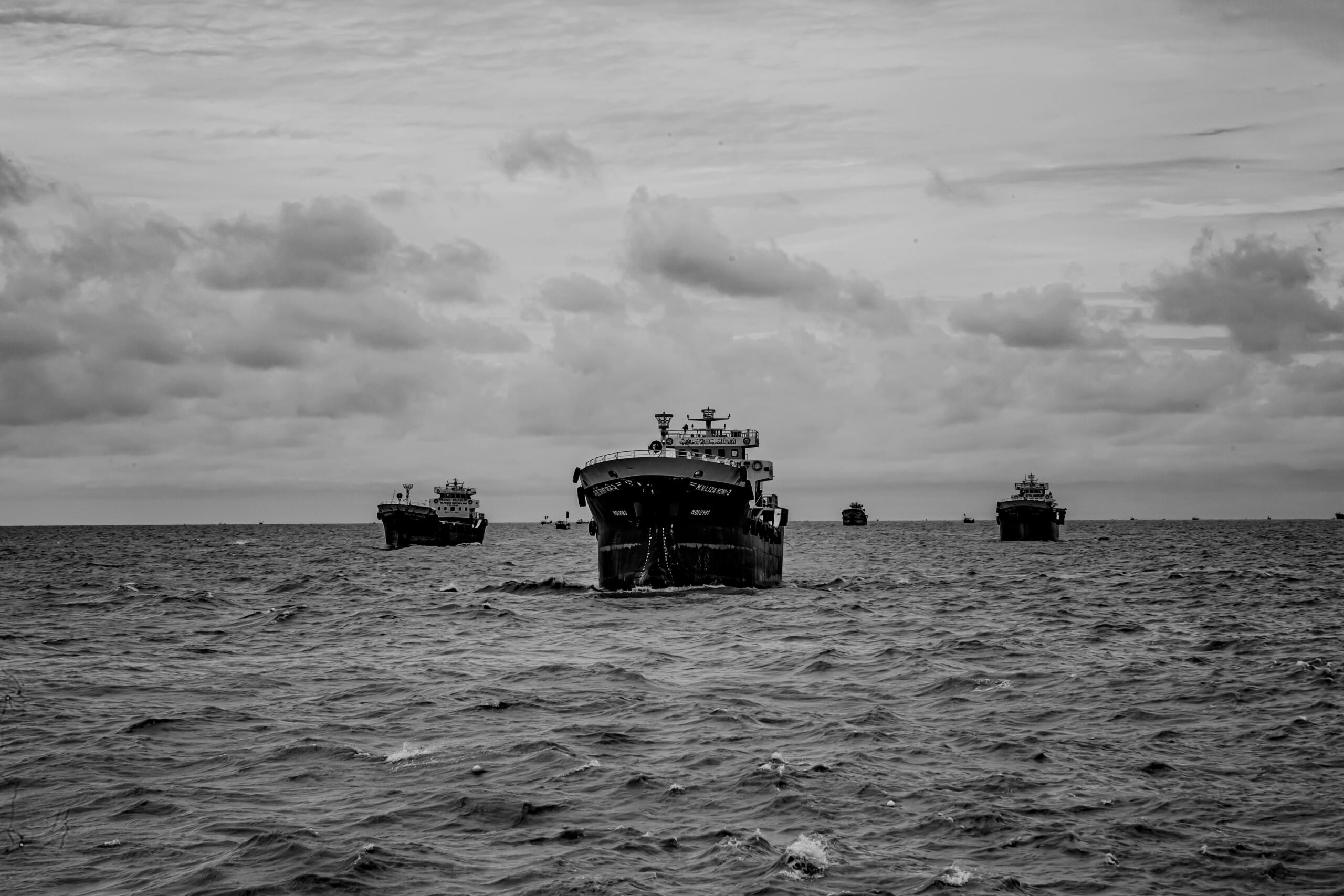 Black-and-white image of several cargo ships sailing in a line across a rough sea under a cloudy sky.