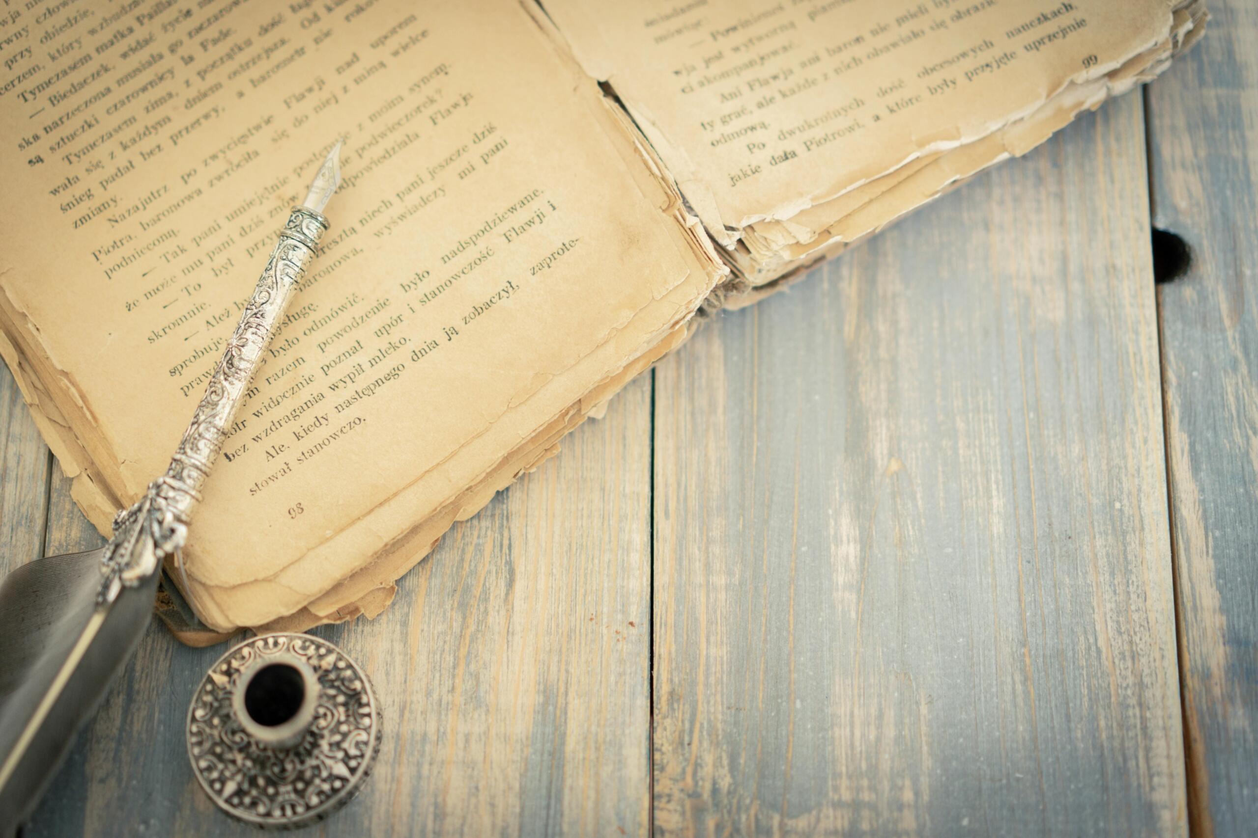 Silver quill and ink pot resting on an old book page on a wooden table.