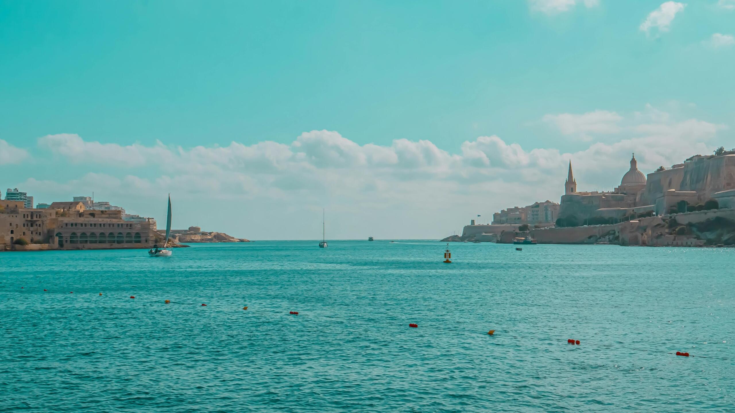 Wide view of Malta’s Grand Harbour with turquoise water, small boats, and historic fortifications and domes along the shoreline under a bright sky.