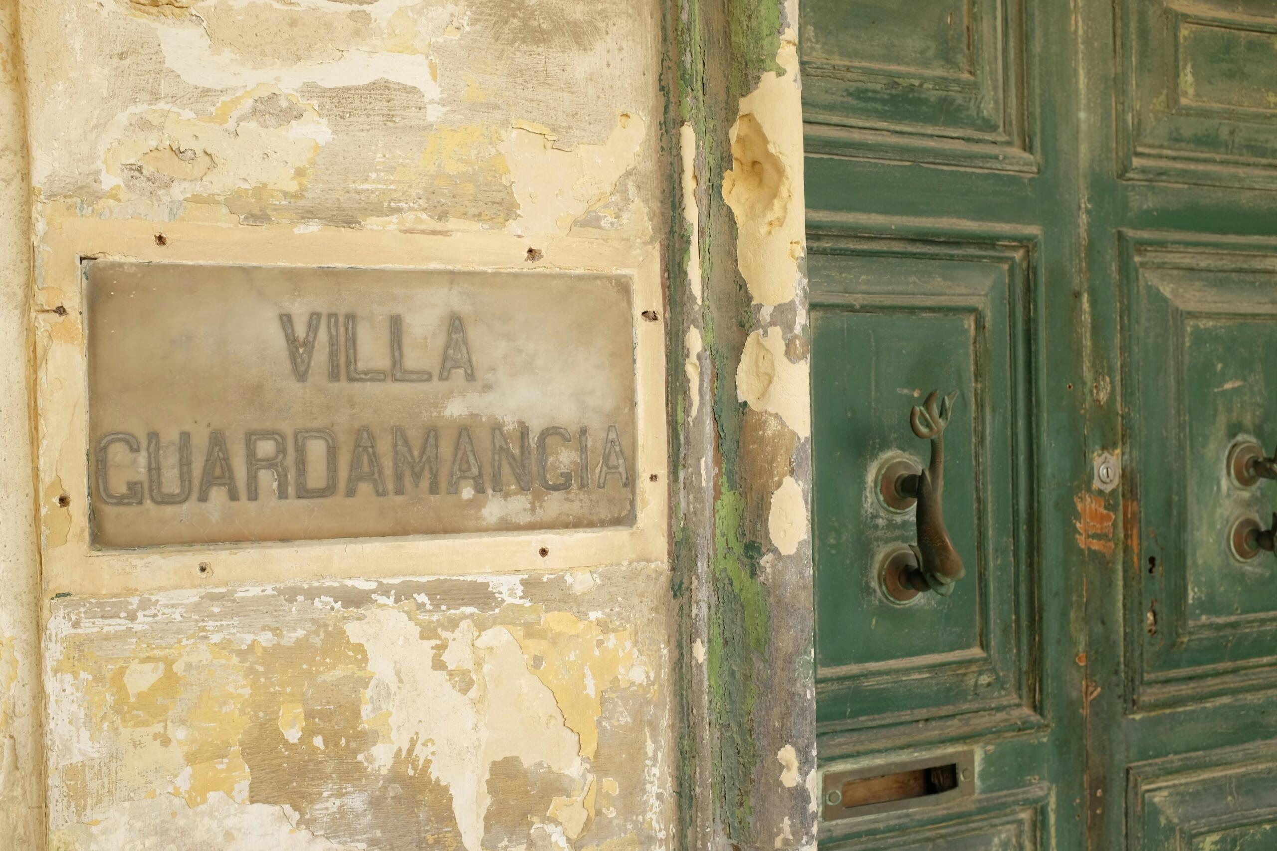 A weathered limestone wall with peeling paint frames a plaque reading “Villa Guardamangia” beside a green wooden door with a metal handle.