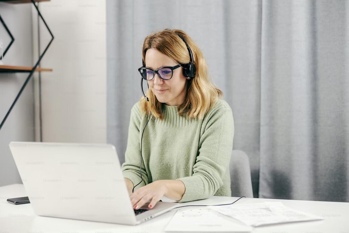 A woman wearing glasses and a headset, sitting at a desk and working on a laptop with papers beside her, suggesting an online French lesson or remote language study session.