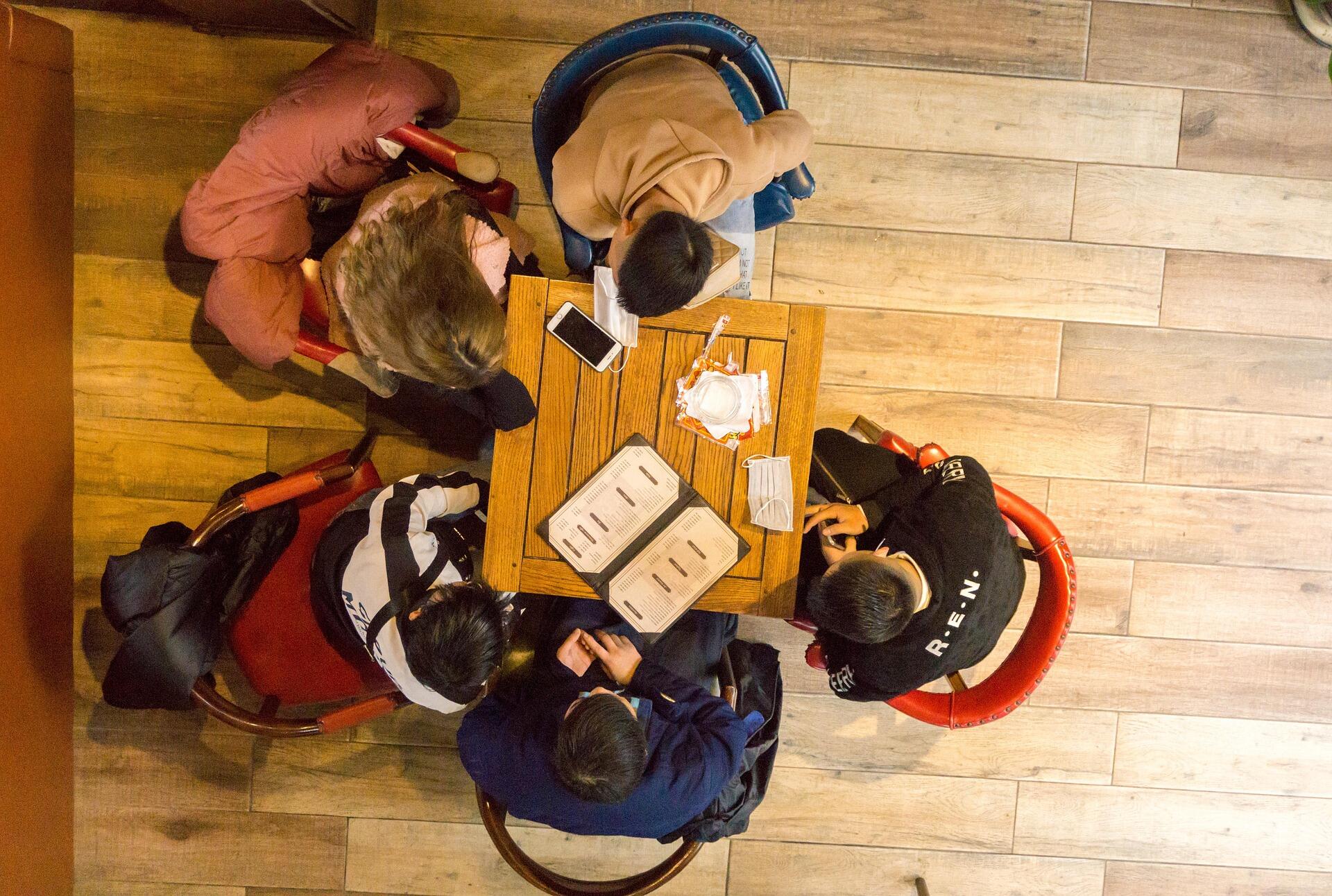 Top-down view of five young adults sitting around a small wooden café table, looking at menus and a smartphone, with face masks placed on the table and wooden flooring visible around them.