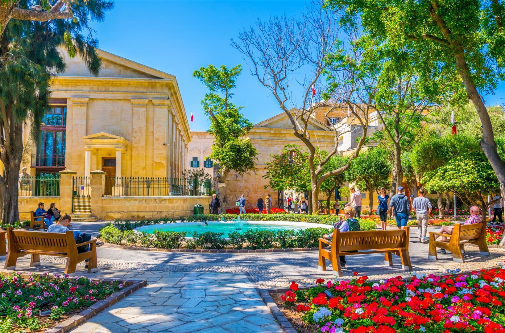 A sunny day at Upper Barrakka Gardens in Valletta, Malta, showing people relaxing on benches near a fountain and colorful flower beds with historic limestone buildings in the background.