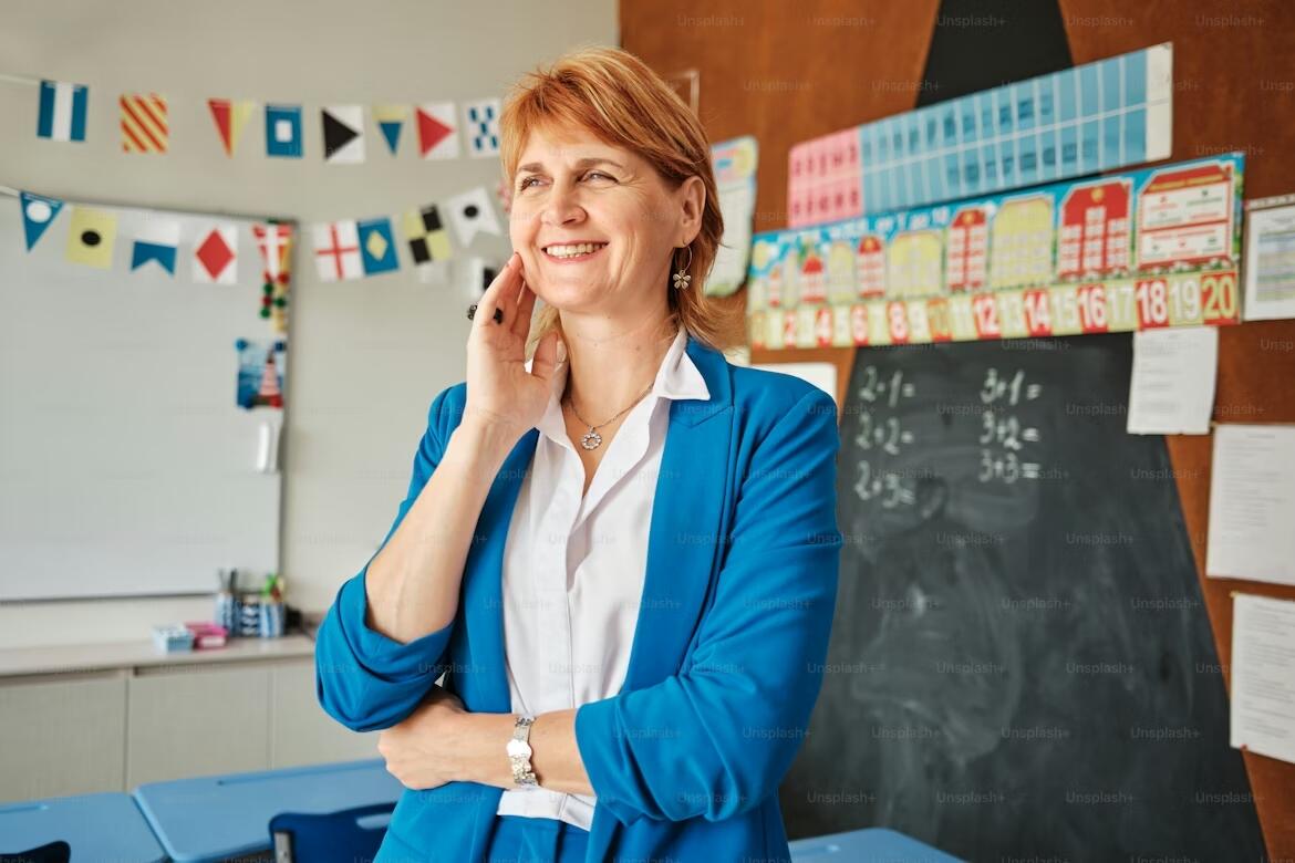 Confident French teacher standing in a bright classroom with a chalkboard and colorful learning materials in the background, smiling while preparing for a lesson.