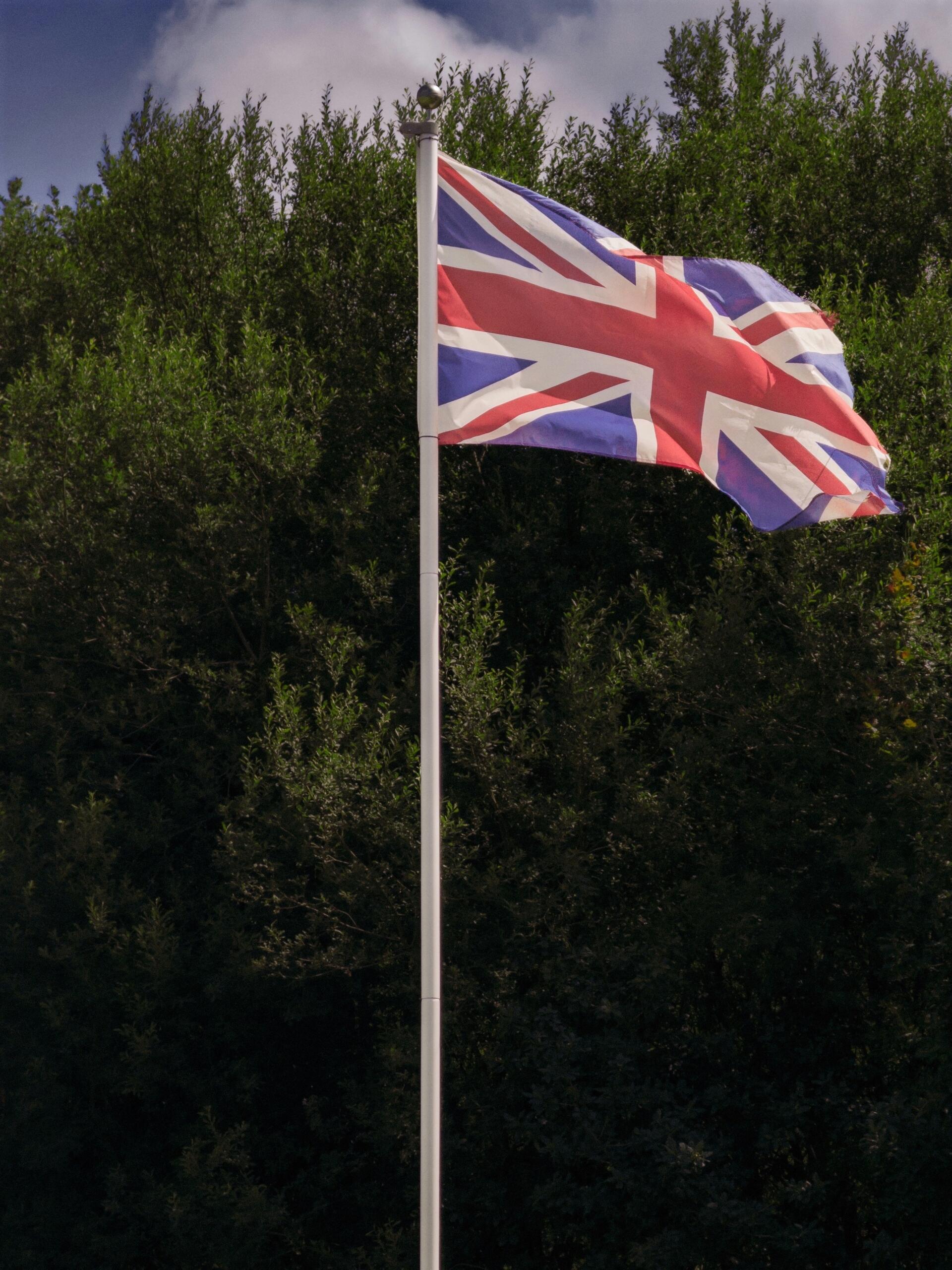 The Union Jack flag waves in front of leafy green trees against a cloudy sky.