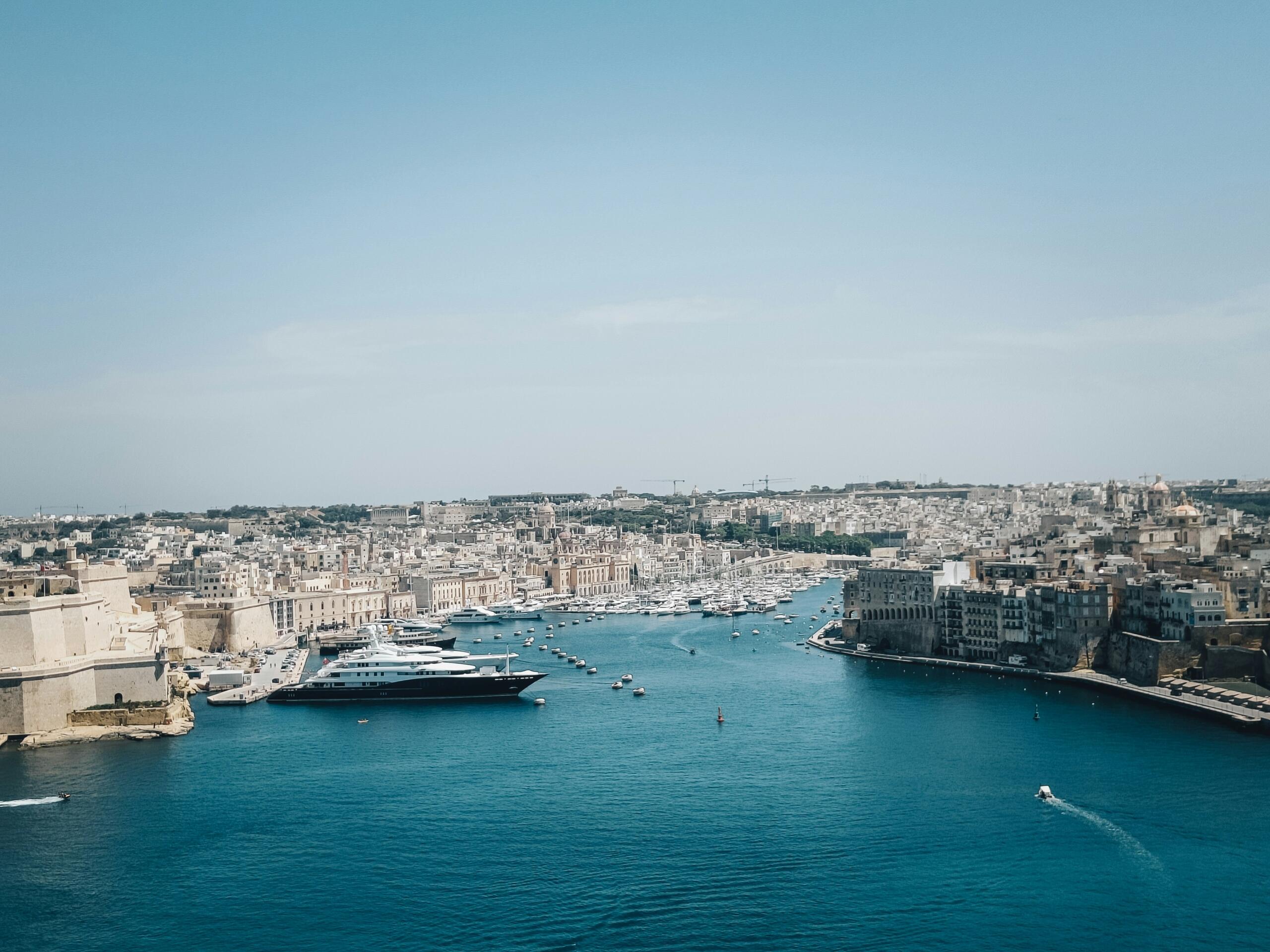 A cityscape of Valletta, Malta, with historic buildings lining the waterfront under a blue sky and the sea in the foreground.