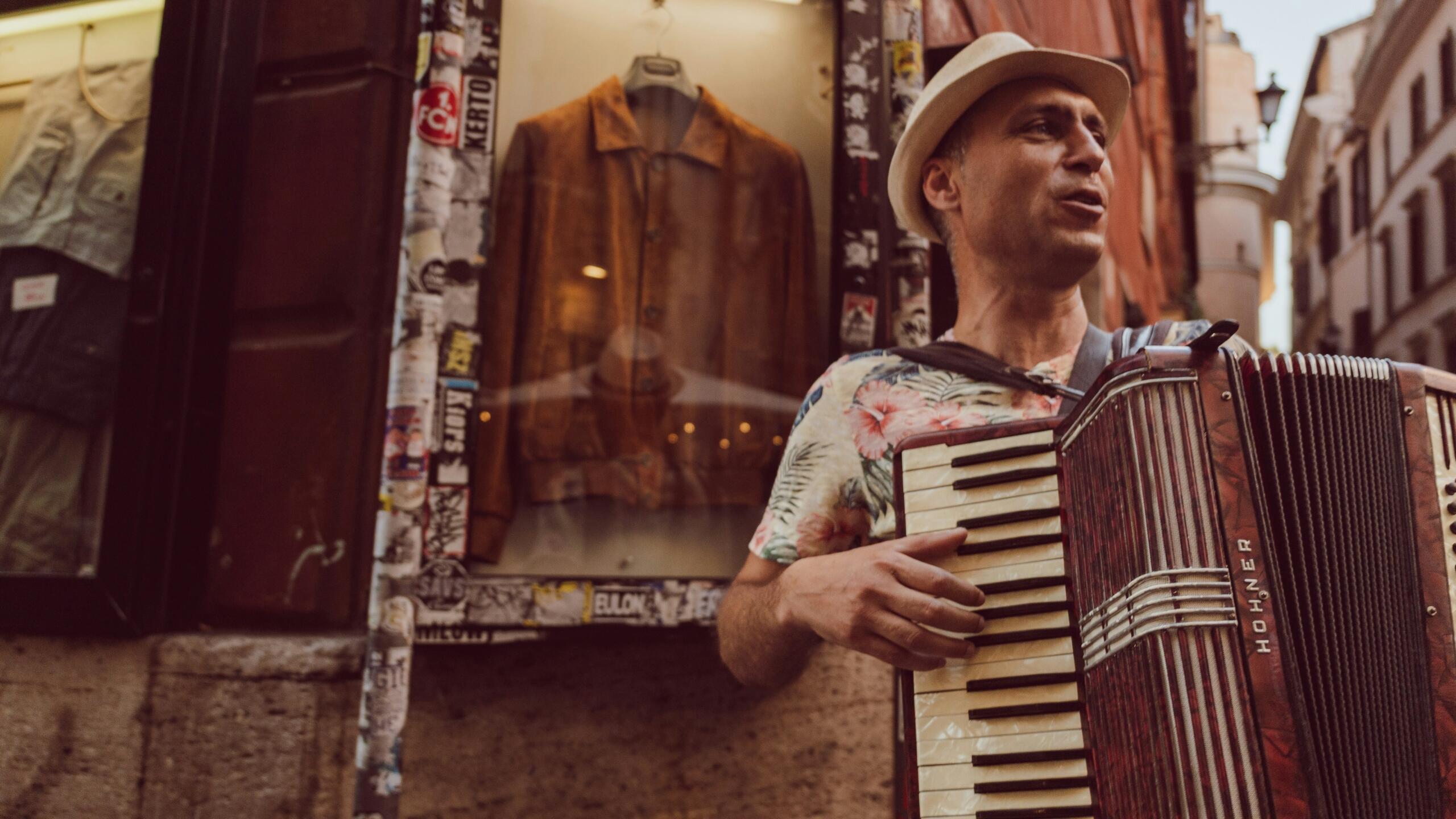 A street musician in Rome plays an accordion outside a shop window, wearing a hat and coat.