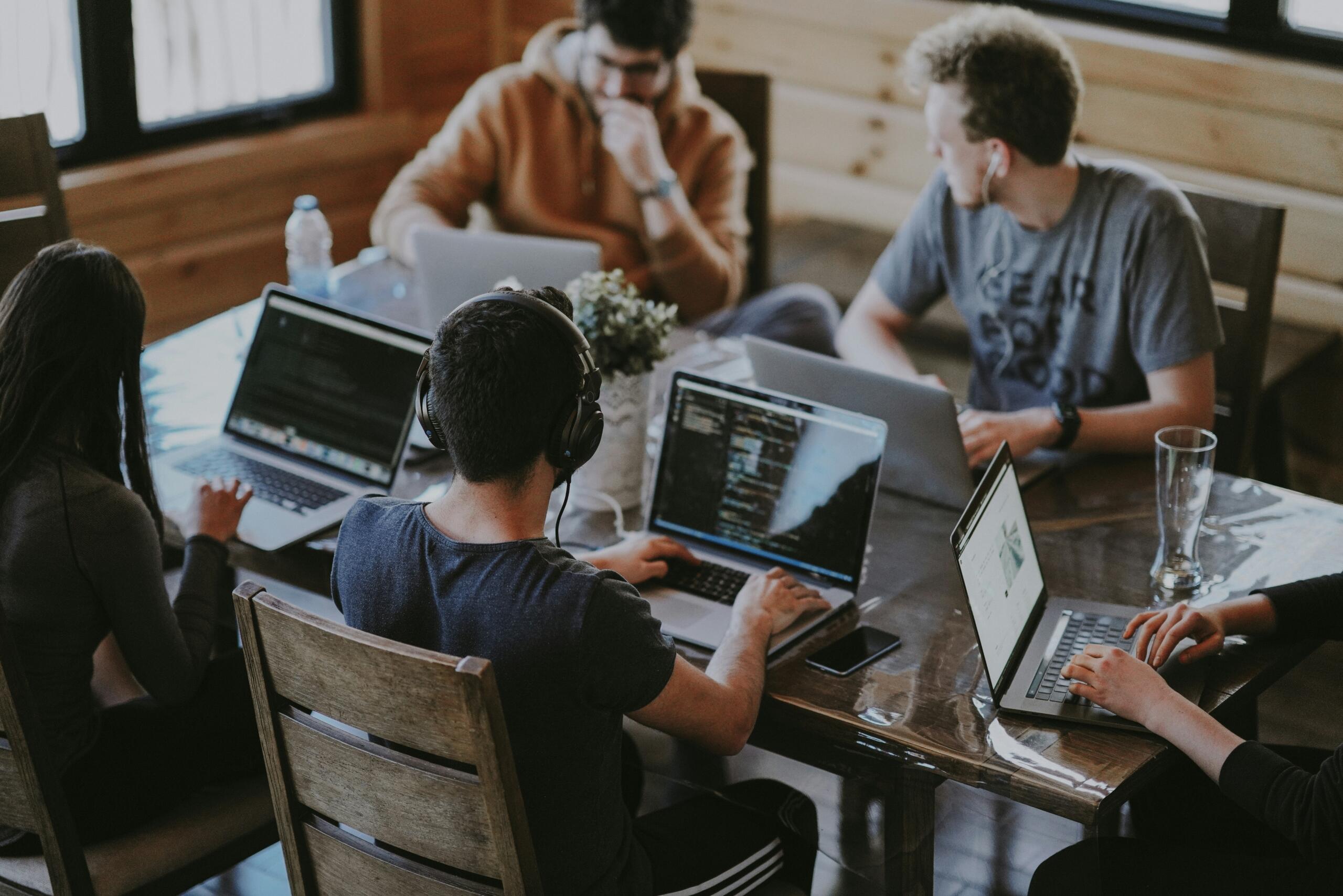 Four young people sitting around a wooden table with laptops and notebooks in a cosy room, working together on a project.