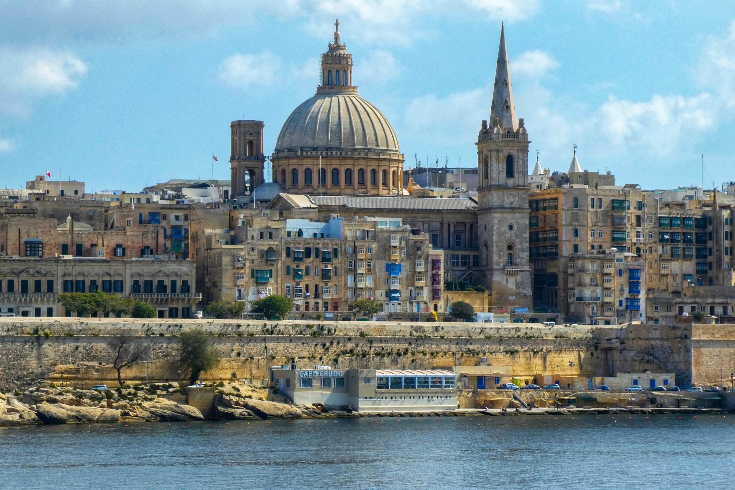 Panoramic view of Valletta’s skyline with the dome of St Paul’s Pro-Cathedral and a tall spire, seen across the water under a blue sky.