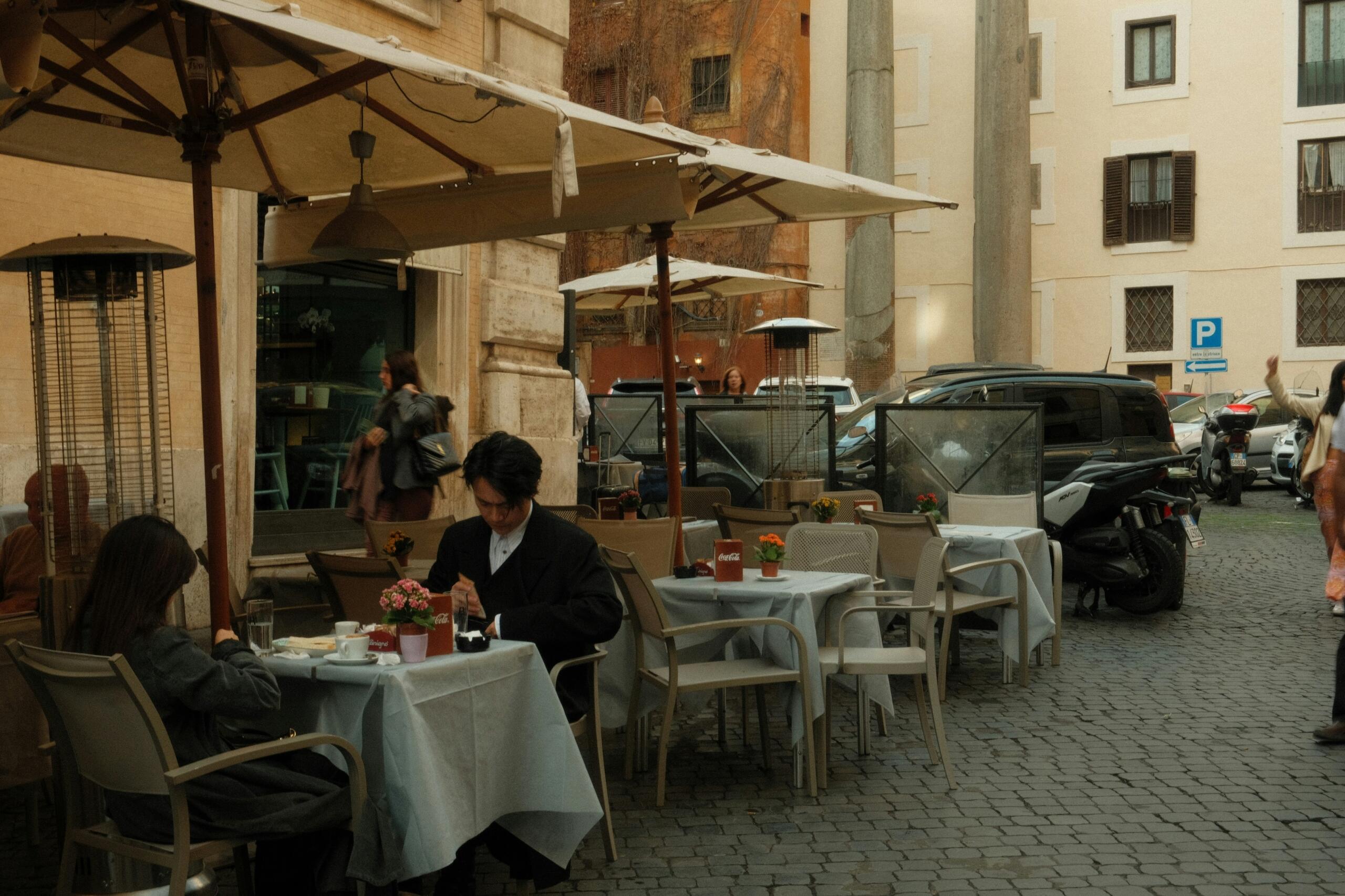 An outdoor café in Rome with tables and chairs under large umbrellas, where people sit and enjoy drinks on a cobbled street.