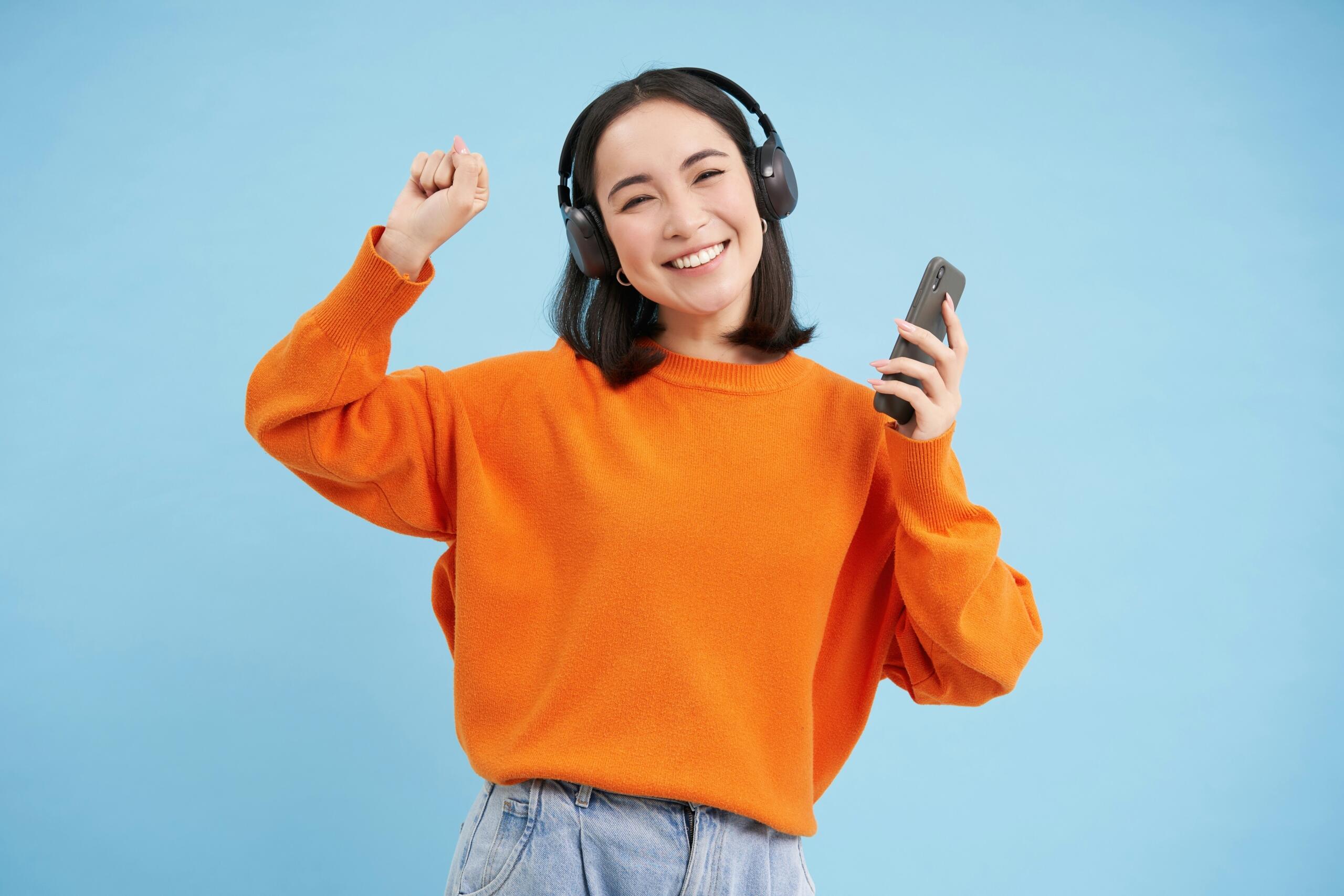 A smiling woman wearing headphones holds a smartphone and dances against a blue background.