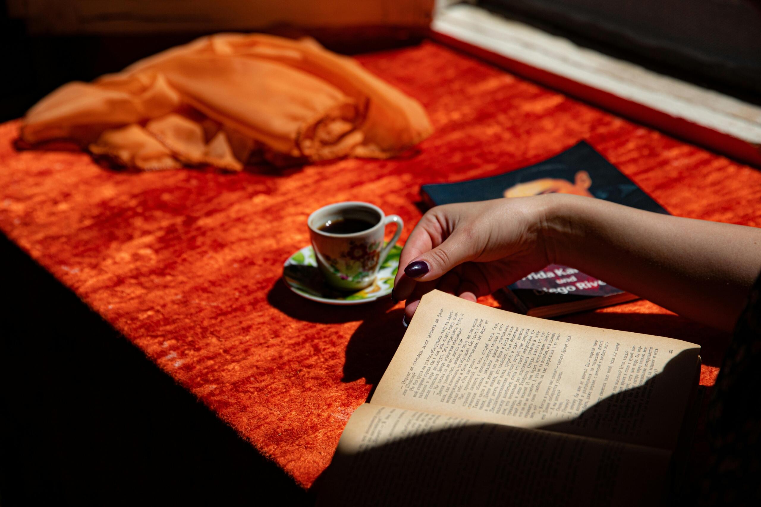 A hand turns pages of an open book on a bright orange table next to a floral coffee cup and a scarf.