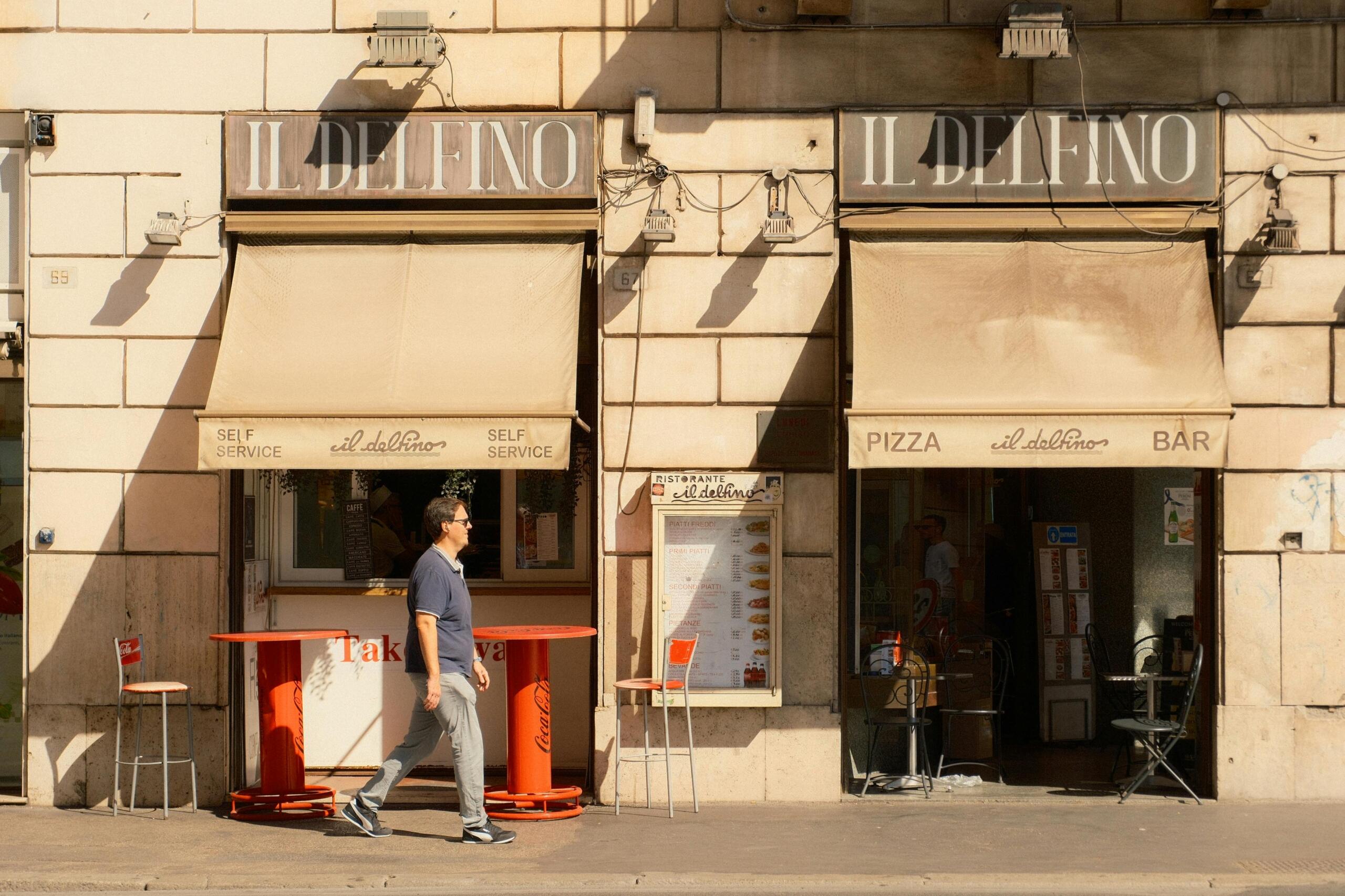 A man walks past Il Delfino café with beige awnings and outdoor tables on a sunlit street.
