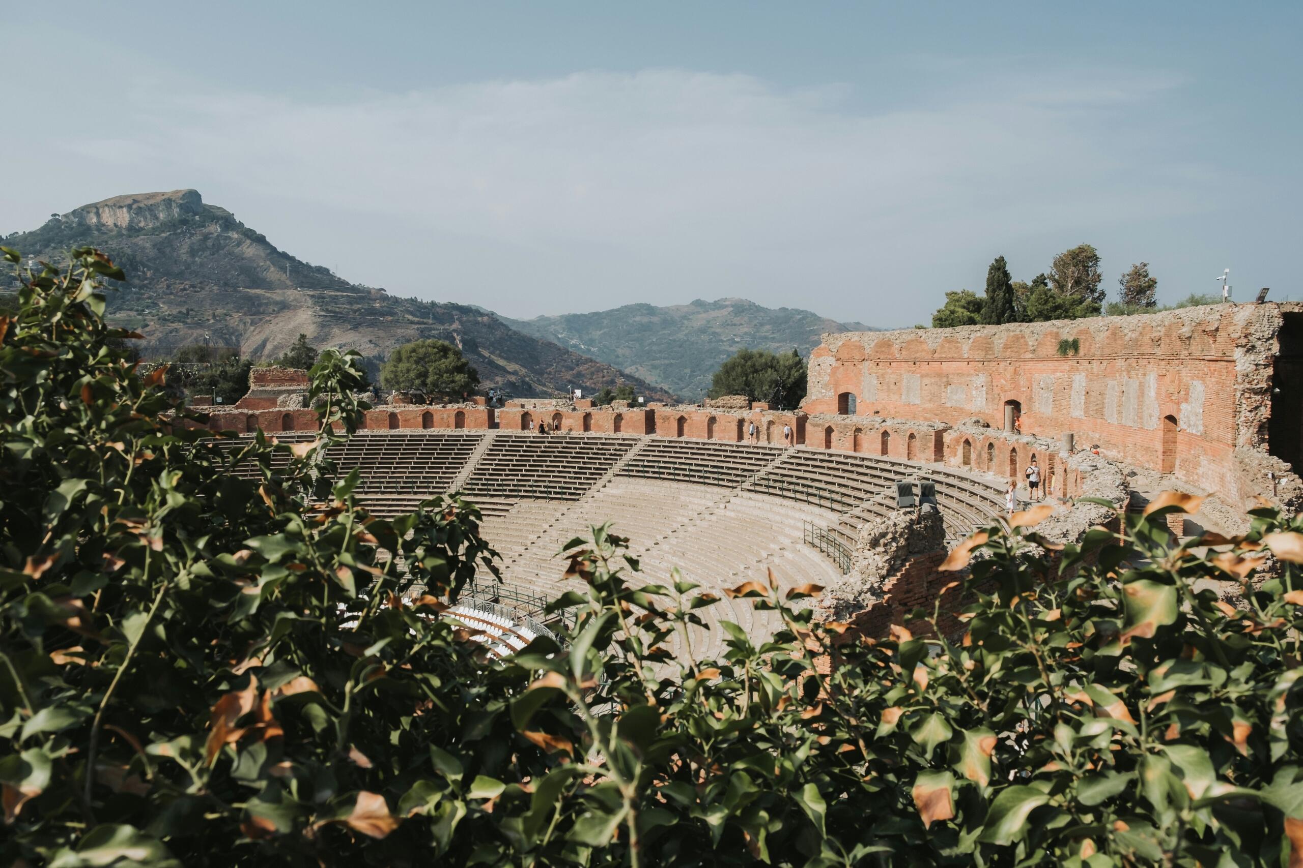 Ancient Roman amphitheatre with mountain views in the background, framed by green foliage