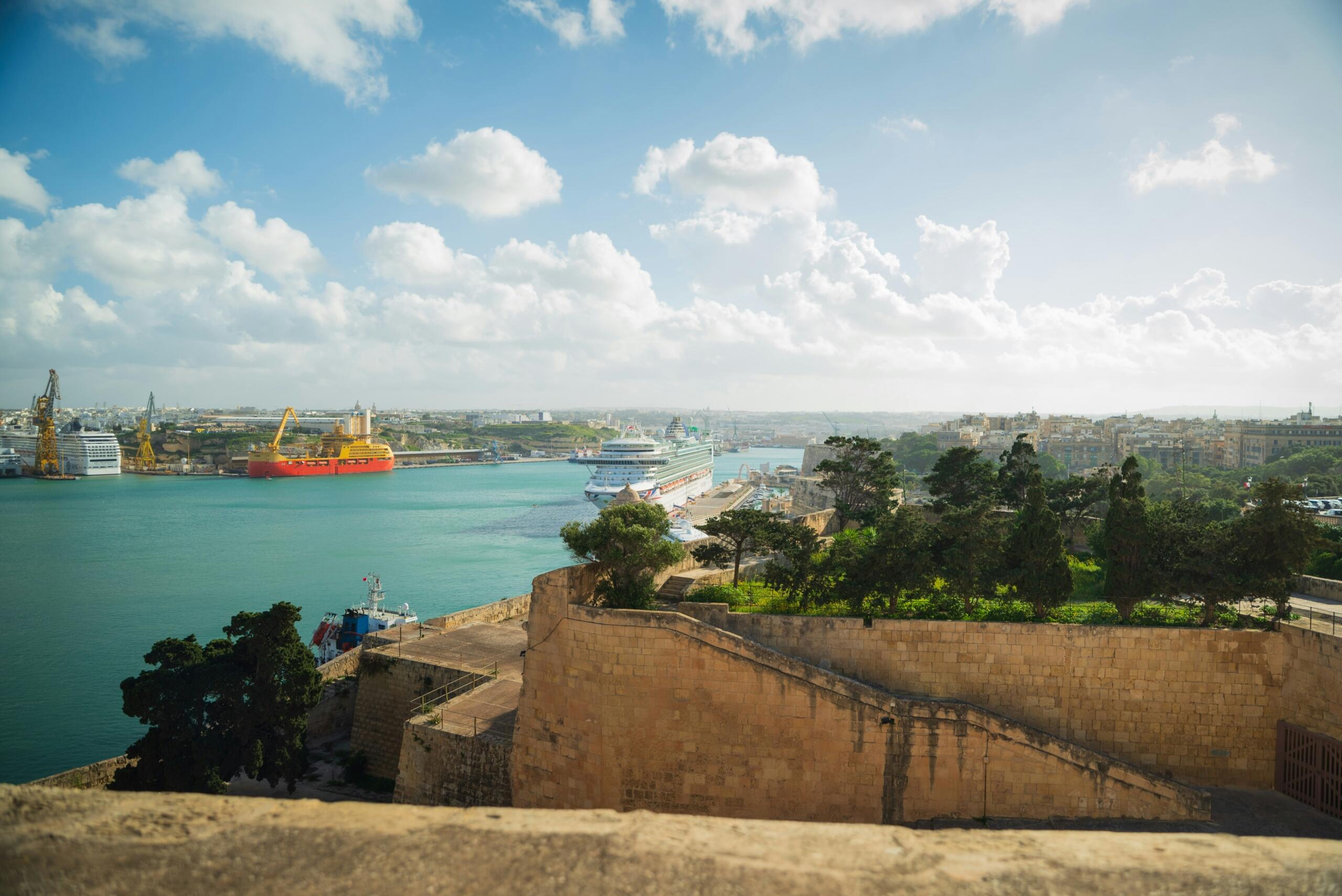 View over Valletta’s harbour with cruise ships, cargo cranes and the city skyline beneath a bright, partly cloudy sky.