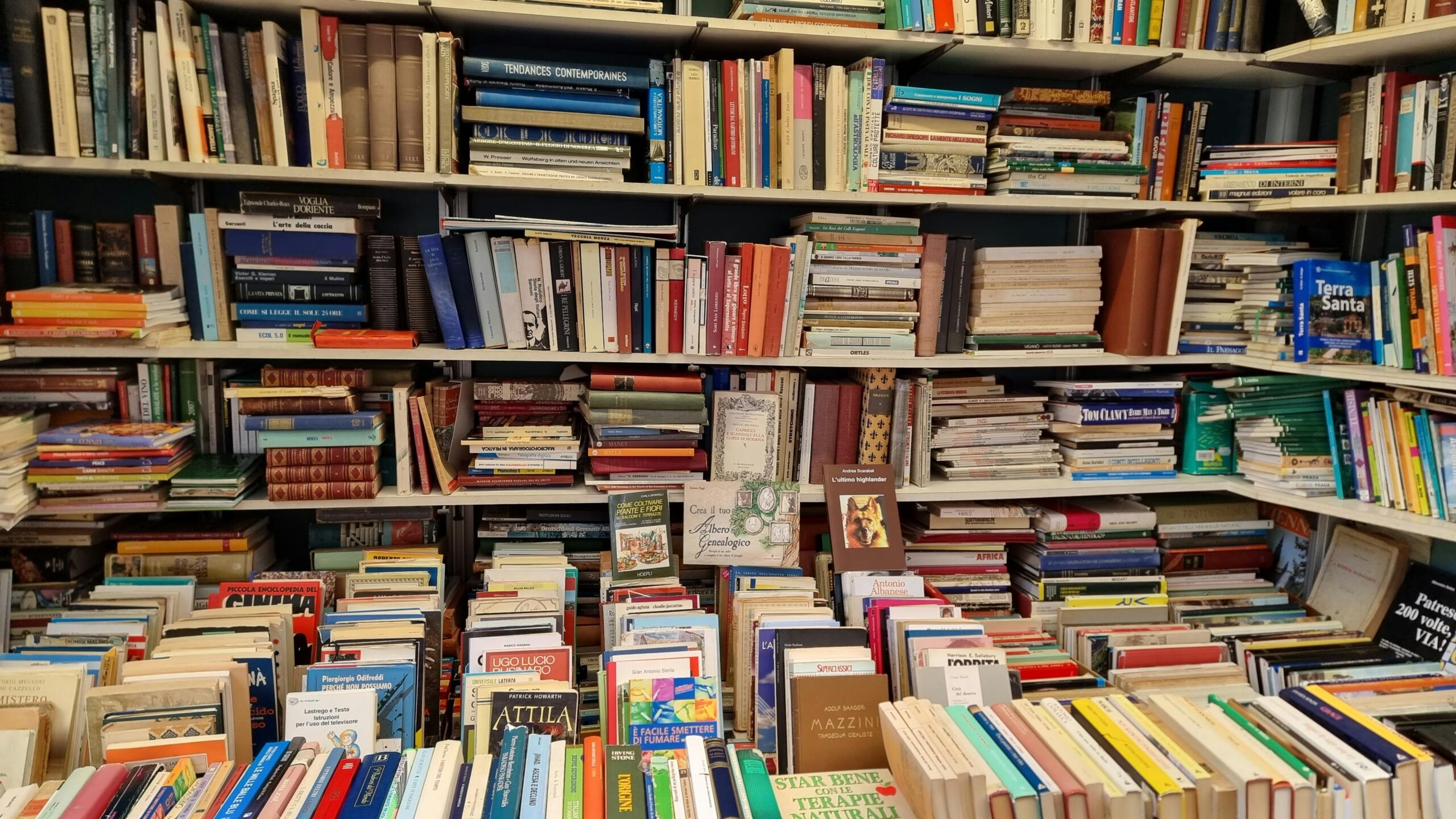 A densely packed bookshelf in an old Italian bookstore with stacks of books on both shelves and tables.