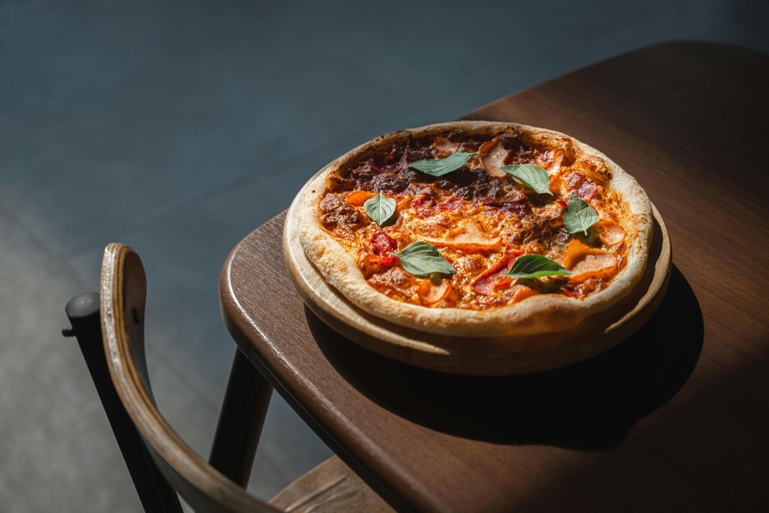 A freshly baked pizza topped with basil sits on a wooden table, lit by a shaft of sunlight.
