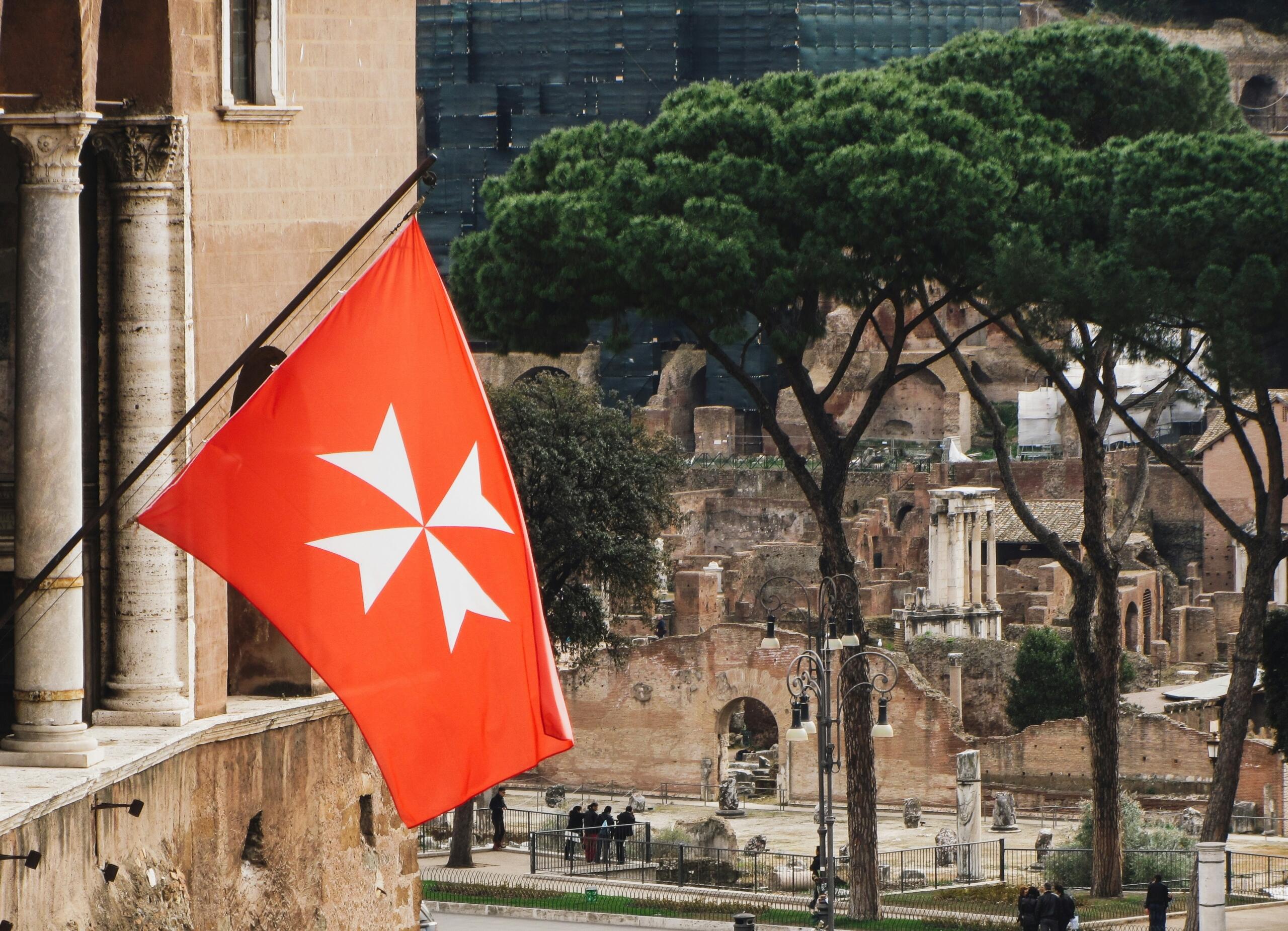 Red Maltese cross flag at Villa del Priorato di Malta in Rome with historic ruins and trees in the background