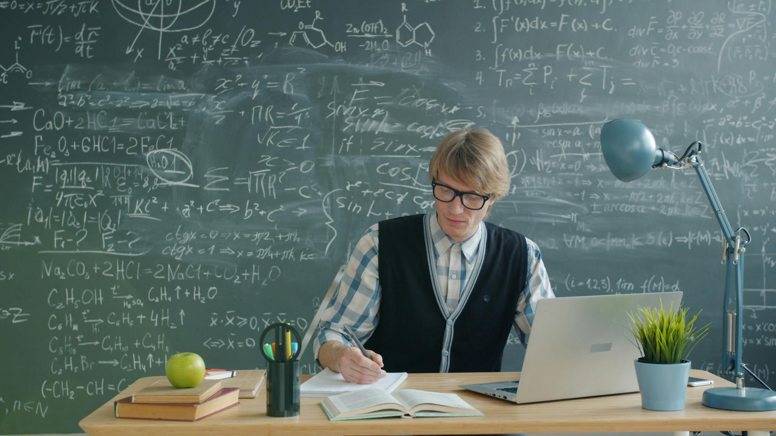 A student sits at a desk with a laptop in front of a chalkboard filled with mathematical formulas.