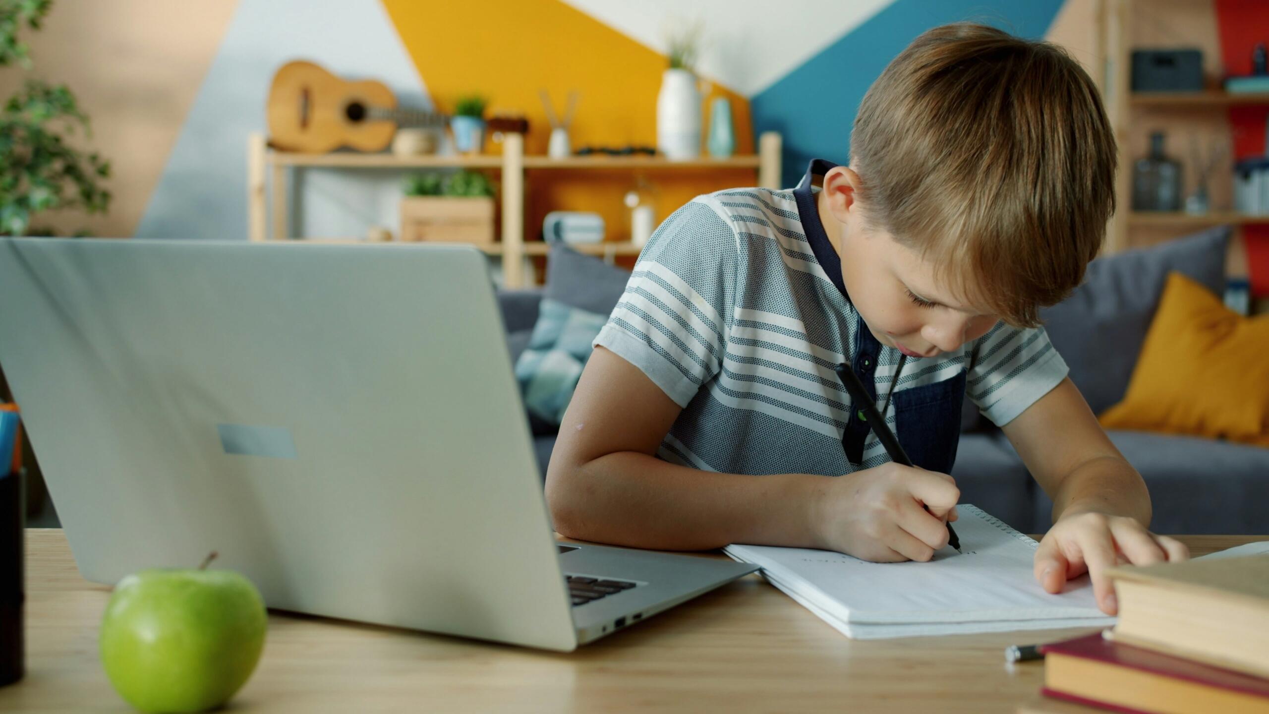 A boy sits at a desk with a laptop and writes in a notebook.