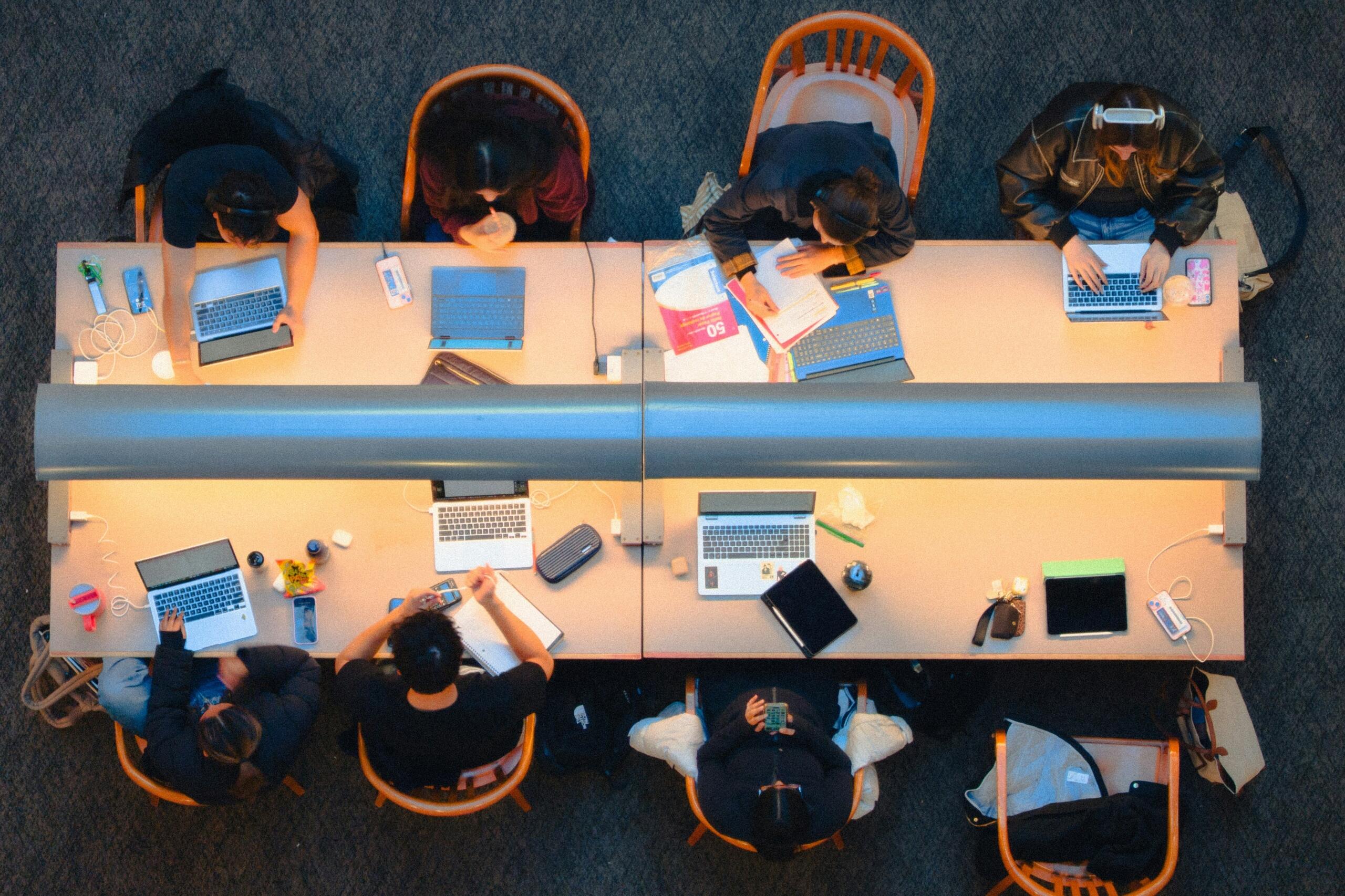Students sit together at a long table with laptops, notebooks, and study materials.