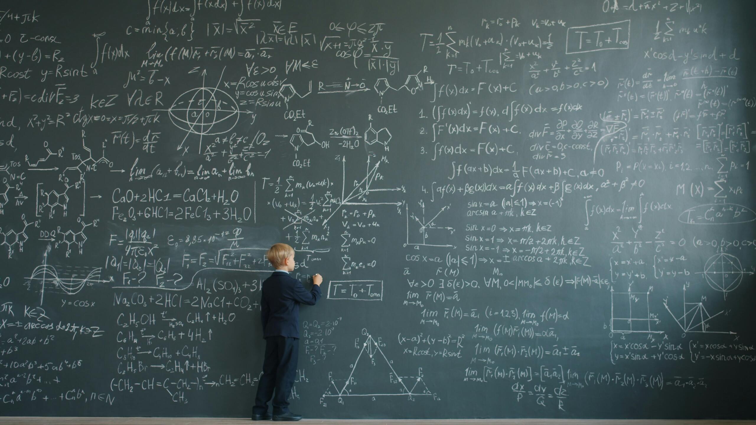 A young boy writes formulas on a large chalkboard in a classroom.