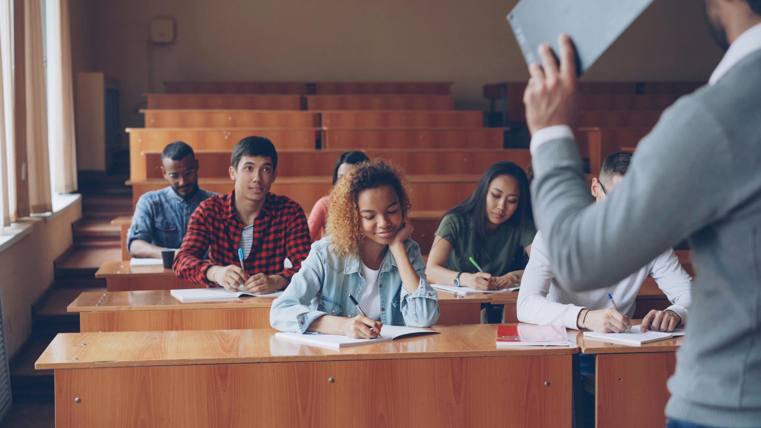 Students sit at tables and write while a teacher speaks in a classroom.