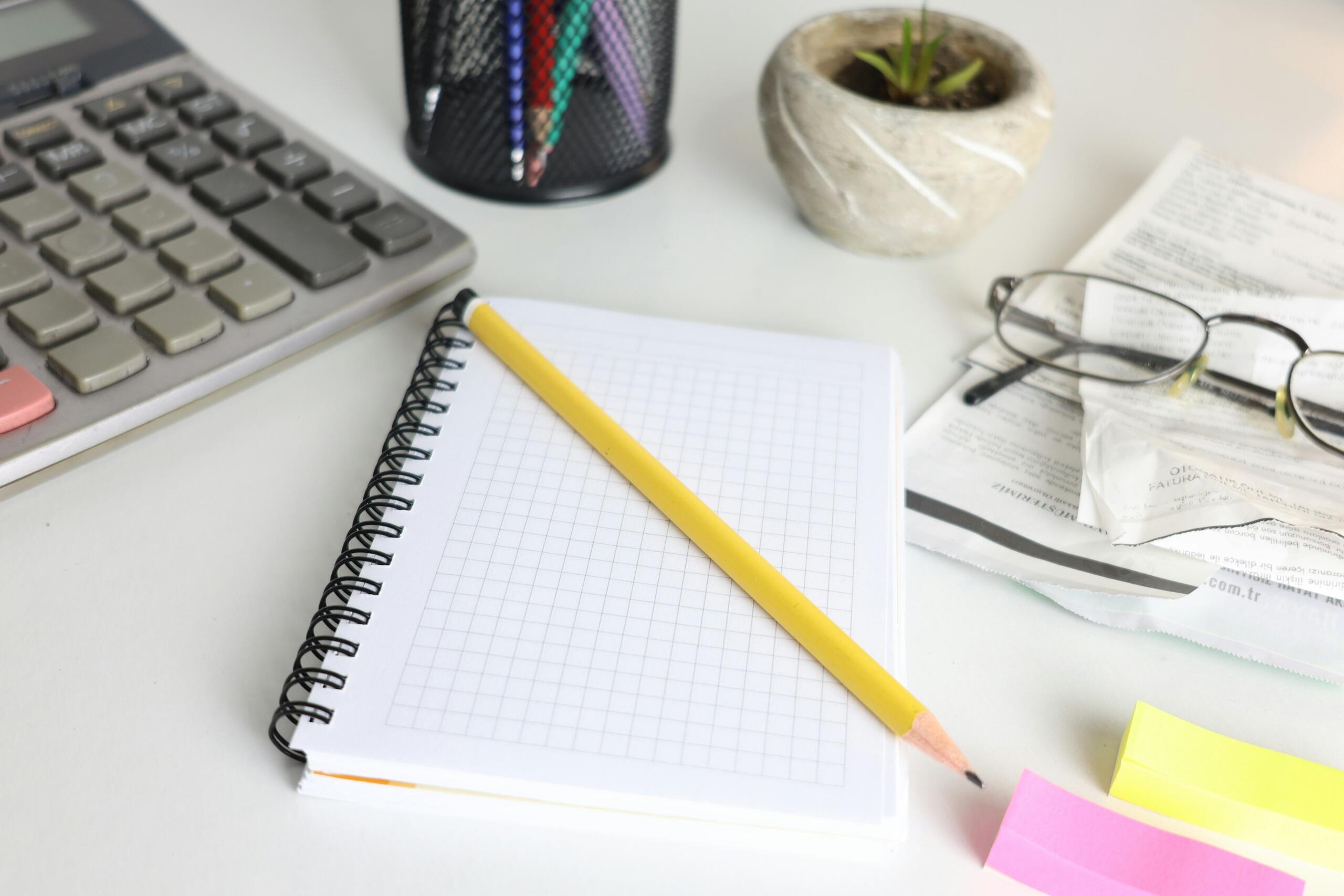 A calculator, notebook, pencil, and glasses lie on a desk.
