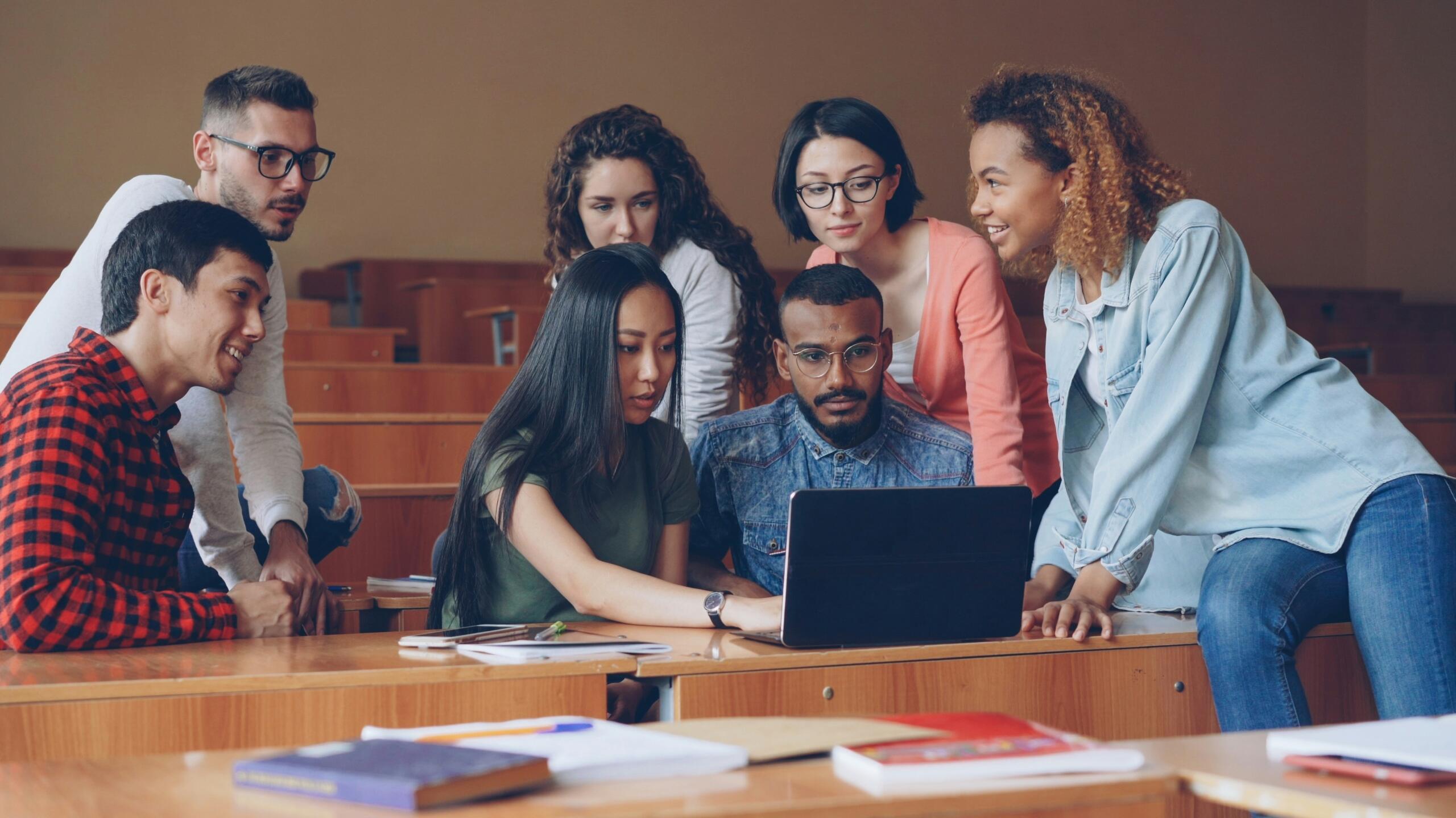 A group of students sit together around a laptop in a lecture hall.
