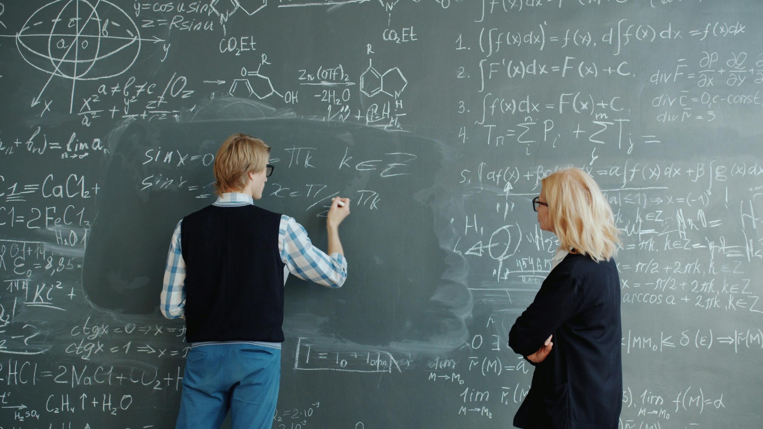 A student writes formulas on a large chalkboard while a teacher watches.