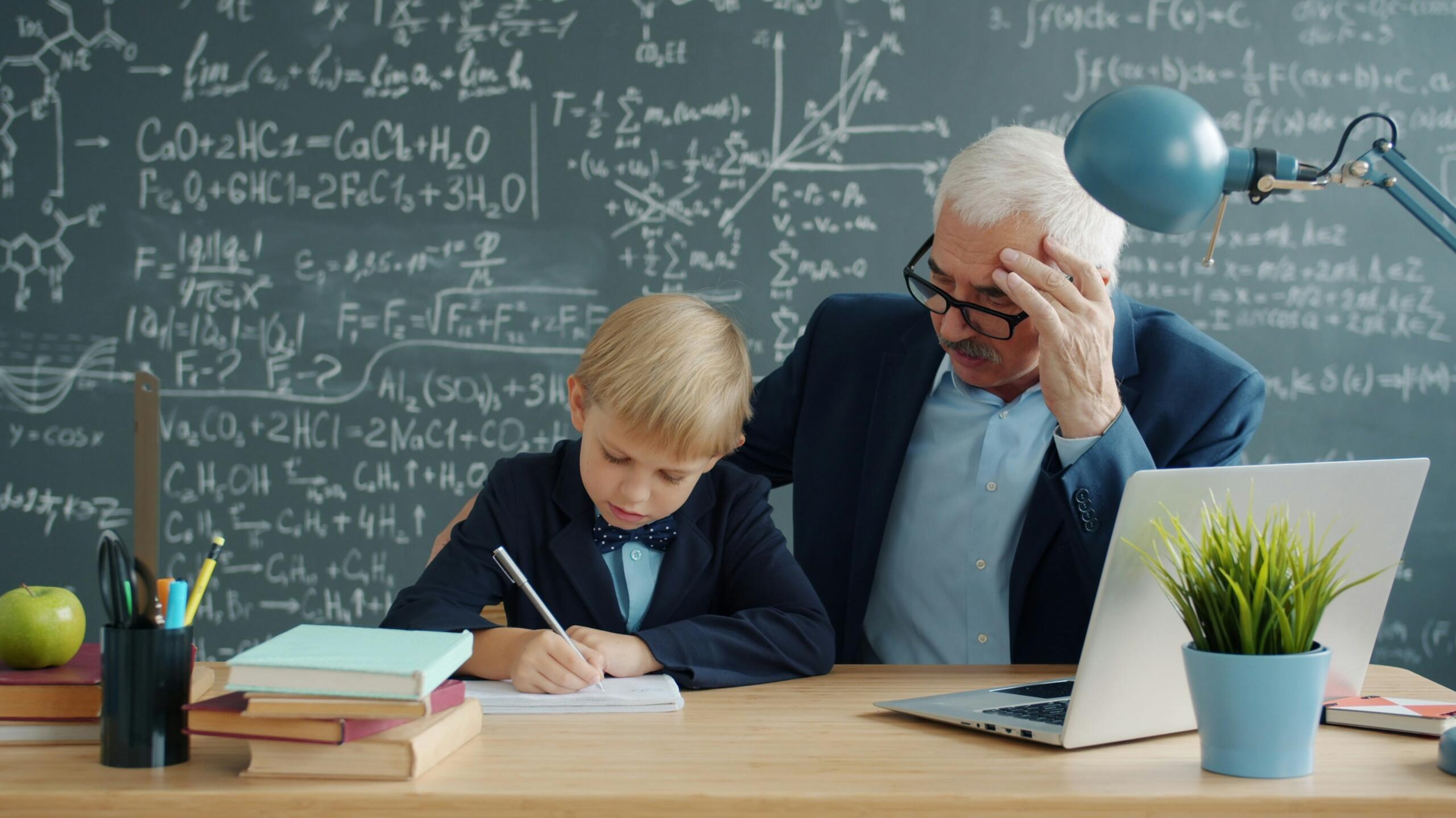 An older teacher sits beside a young student who is writing in a notebook, with a laptop on the desk.